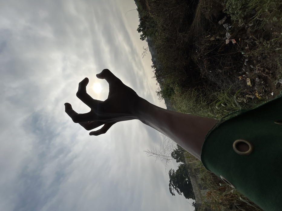 An outdoor, backlit photograph set in Debasco, Nigeria, features a human arm extending from the lower-left foreground. The hand, silhouetted against a partially cloudy sky, is curled with the thumb and forefinger forming a circle, framing the bright, obscured sun. A portion of dark green fabric with a visible brass eyelet is present at the bottom left of the frame. The background consists of dense, overgrown vegetation and scattered refuse on the right, while the left horizon reveals distant trees and partially constructed concrete block structures under a largely overcast sky.