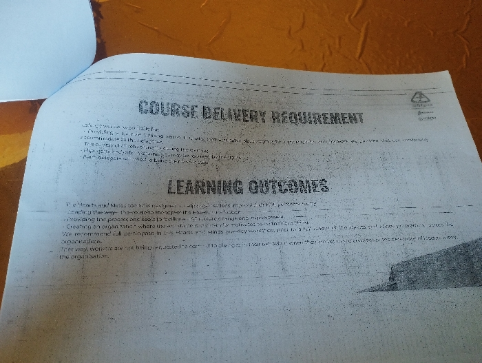 The image shows a page from a printed document, likely a manual or training guide. The document’s header reads “COURSE DELIVERY REQUIREMENT” and “LEARNING OUTCOMES” The text within the document is illegible due to poor image quality. The image is taken from a location on the planet Earth with coordinates 10.917 latitude, 11.476 longitude, which corresponds to the city of Ashaka, Nigeria. The image is taken on a surface that appears to be a desk, possibly in an office or educational setting. The image is taken at a slight angle from above the surface, and the light source is unknown.