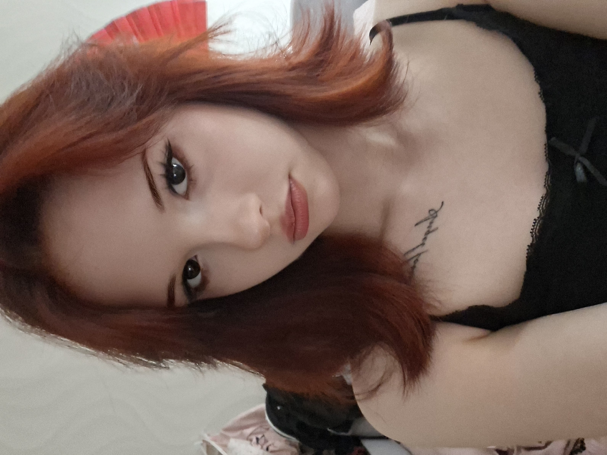 A young woman with reddish-brown hair is positioned in a close-up, low-angle shot. She has pale skin, brown eyes with visible makeup, and light red lipstick. She is wearing a black lace top. A small tattoo is visible on her upper chest. The background is blurry and indistinct, but includes elements suggesting an interior setting with white and patterned surfaces. The image provides no clear indication of the location of Chaikovsky, Russia.