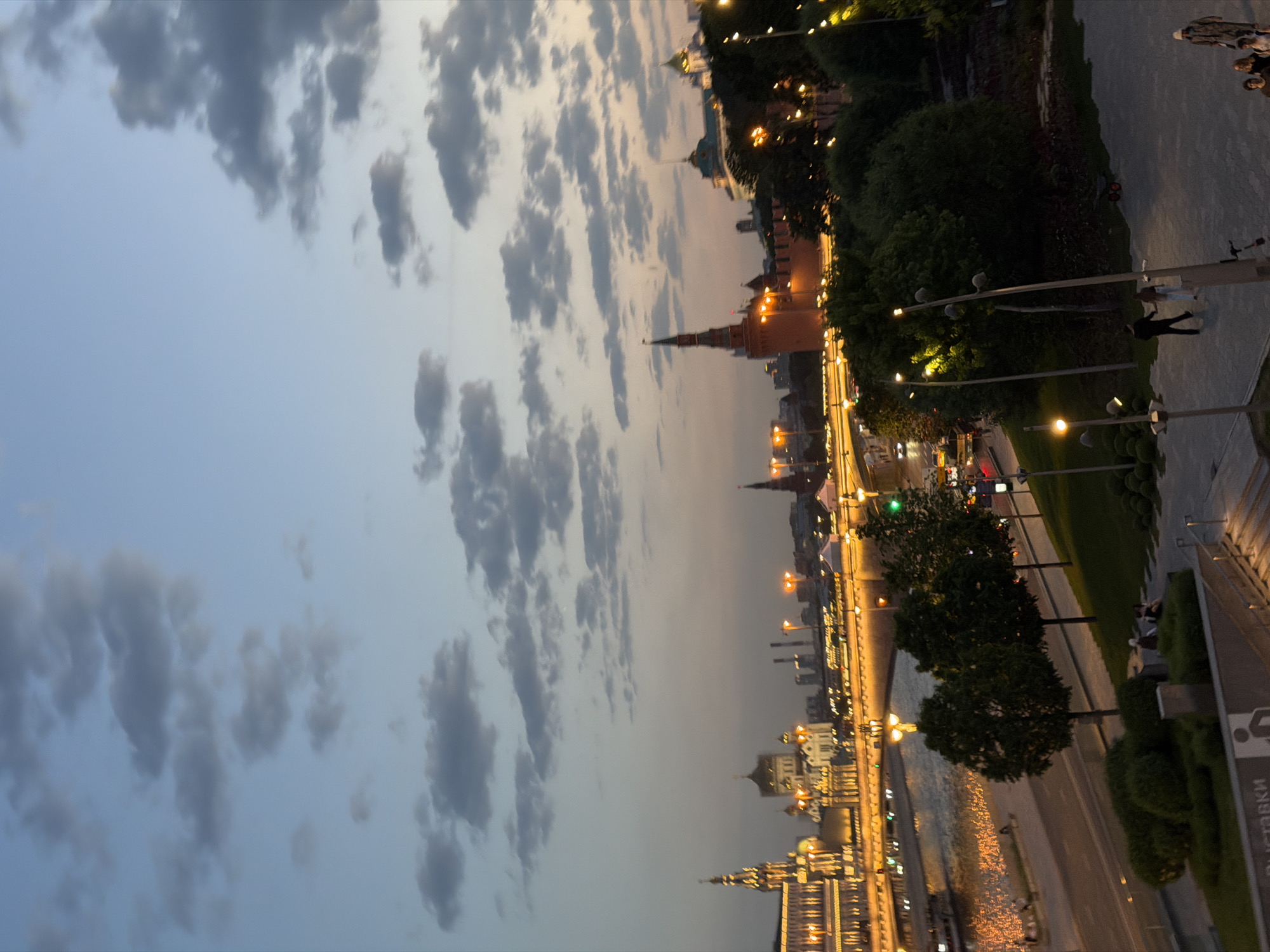 The image shows a cityscape at dusk. In the foreground is a sidewalk, with lampposts and trees. The background features the Moscow Kremlin, with its distinctive towers and golden lights. A river runs parallel to the embankment. The sky is filled with grey clouds. People are walking along the sidewalk.
