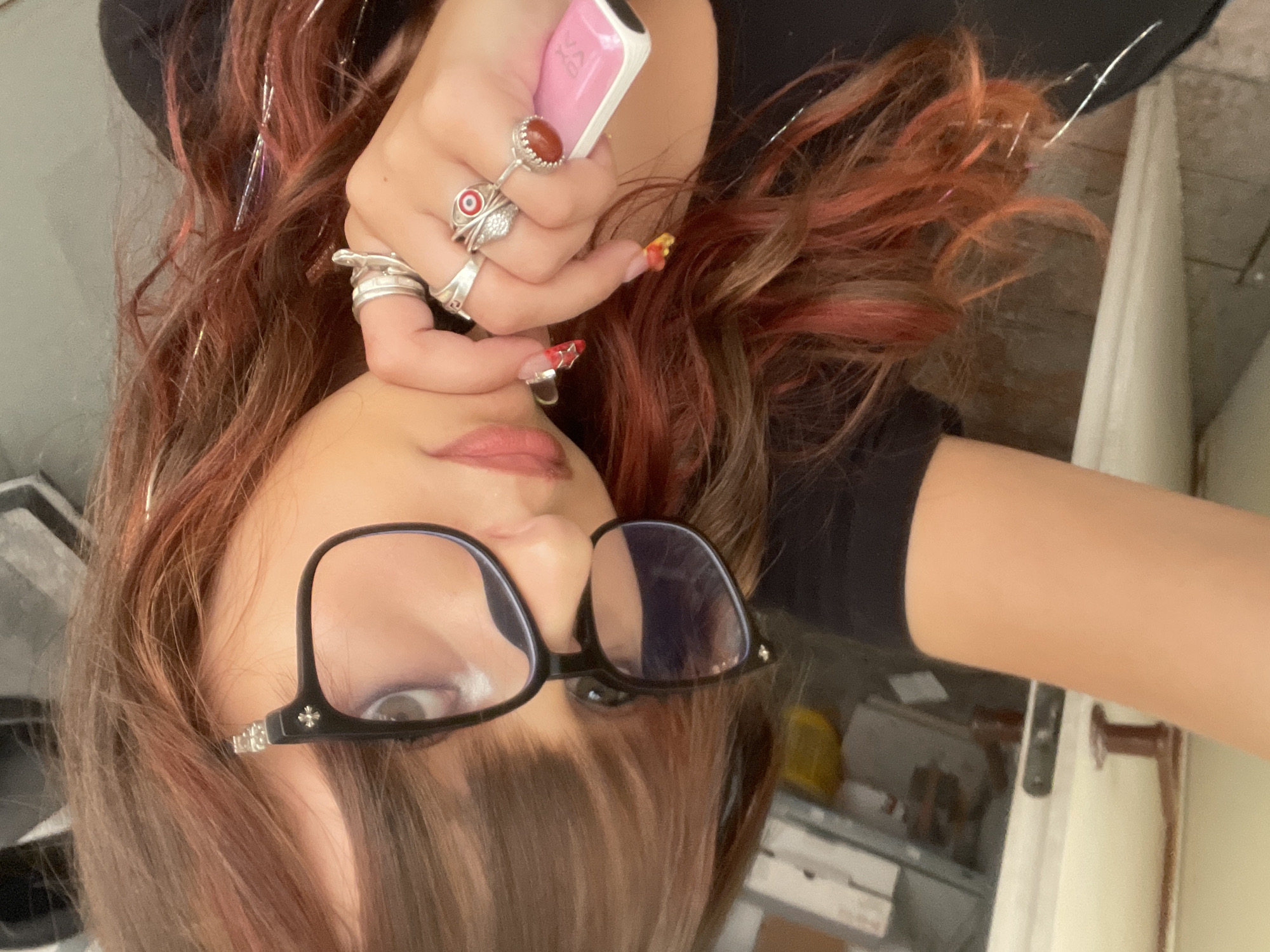 In a selfie taken at a close range, a person with reddish-brown hair adorned with silver tinsel is seen. They are wearing black-rimmed glasses with subtle decorative elements on the temples. The person is also wearing several silver rings on their fingers, including one with a red evil eye design and another with an orange-colored stone. A pink electronic device is held in their right hand. The background appears to be an indoor environment with muted colors, possibly a room with furniture and a window. The overall impression is a casual, personal snapshot. There is no discernible indication of the location being Raanana, Israel.