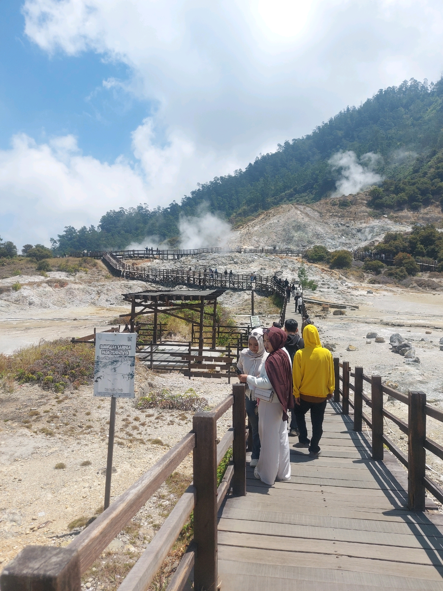 A group of tourists are walking on a wooden boardwalk that winds through a geothermal area in Sikunang, Indonesia. The landscape is characterized by barren, rocky terrain with patches of dry vegetation. Steam rises from various points in the ground, creating a hazy atmosphere. In the background, a forested hillside slopes upward, and the sky is a bright blue with scattered white clouds. A sign with Indonesian text is visible near the boardwalk. Several individuals are seen on the boardwalk, some walking in groups, while others are further up the path. The architecture of the boardwalk features wooden planks and railings, with some sections elevated above the ground. The overall scene depicts a natural attraction that is being visited by tourists.