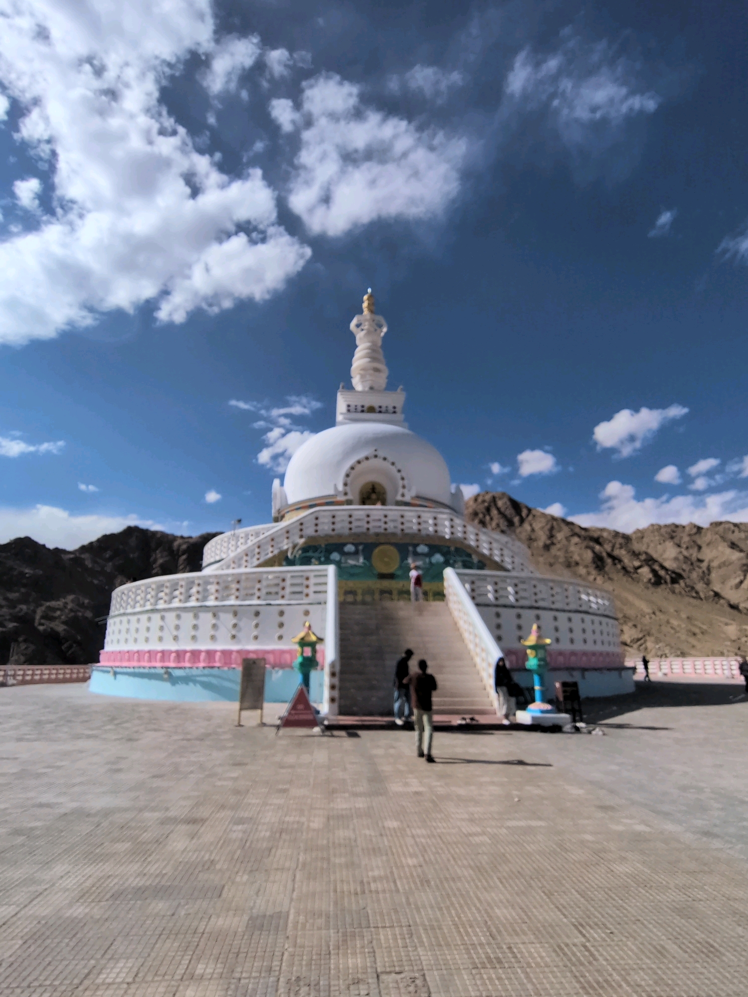 The image shows the Shanti Stupa in Leh, India, a Buddhist white-domed stupa. The stupa is situated on a hilltop with mountains in the background. The sky is blue with white clouds. Several people are visible on the grounds of the stupa, some walking and others standing. The stupa itself is ornate with decorative elements and appears to be a significant tourist attraction. The foreground shows a paved courtyard with a patterned surface.
