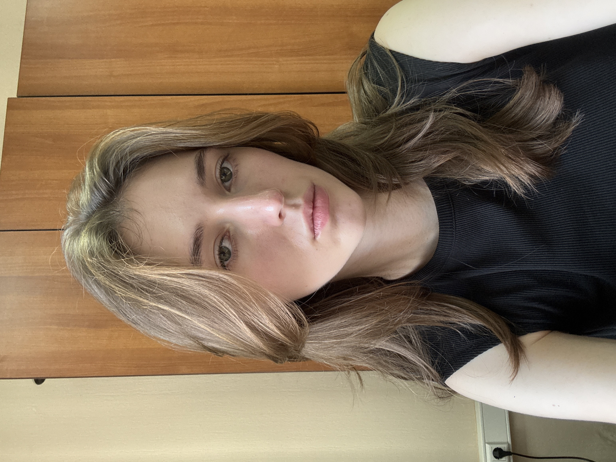 A young woman with shoulder-length, wavy, light brown hair is looking directly at the camera. She is wearing a black sleeveless ribbed top. Her skin appears pale and clear, and her makeup is minimal. She has green eyes and light pink lips. Behind her, there is a wall with wooden paneling. To the left of the wooden paneling, a portion of a pale yellow wall is visible. An electrical outlet with a black plug and cord is mounted on the pale yellow wall, near the bottom left of the frame. The scene appears to be indoors, possibly in an office or residential setting. The lighting is natural and soft, casting no harsh shadows. There are no other people or objects clearly visible in the frame. The image does not provide any specific visual cues to pinpoint a location within the city of Perm, Russia.