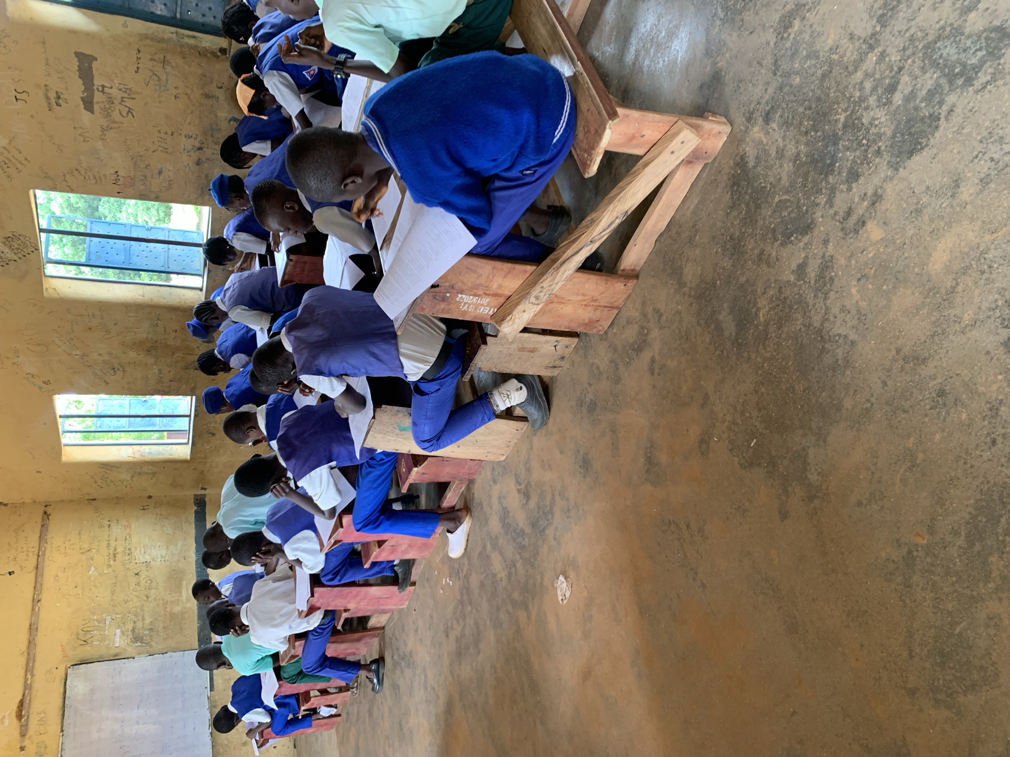 The image depicts a classroom scene in Akwanga, Nigeria, with approximately twenty young students, likely boys, seated at wooden desks. The students are wearing blue and white uniforms, some with blue vests. They are engaged in what appears to be an exam or classwork, as many are looking down at papers. The classroom environment is basic, with a whiteboard on the left wall and two windows on the right wall, one with blue shutters. The walls are yellow and covered in graffiti and markings. The floor is made of concrete and appears to be dusty. The lighting suggests it is daytime. The overall impression is of a humble educational setting.