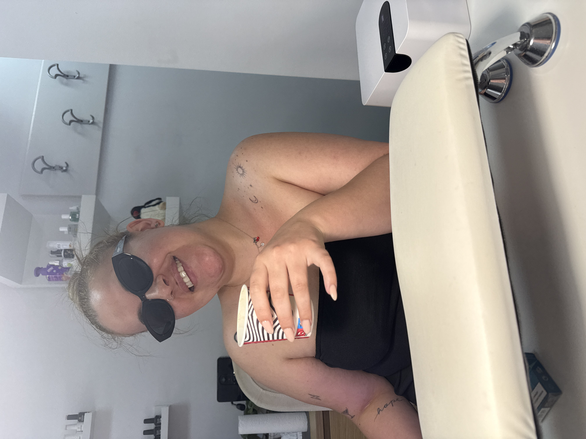 The image depicts a woman with blonde hair in a bun, wearing sunglasses, a black strapless top, and a necklace, holding a disposable coffee cup. She has tattoos on her left arm and shoulder. She is sitting at a white table with a padded armrest in the foreground. To her right, a white nail lamp is visible. The background shows shelves with bottles and a wall with coat hooks. The scene appears to be a beauty salon. There is no indication of the location being Burgas, Bulgaria.