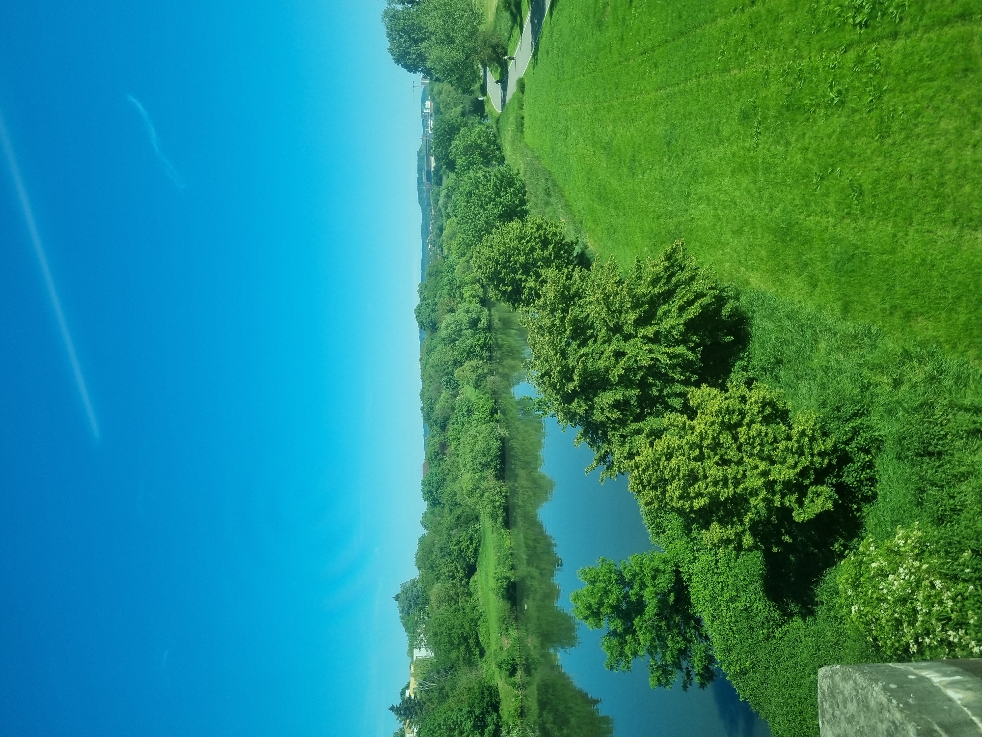 The image depicts a horizontal view of a landscape in Bad Wimpfen, Germany. The top portion shows a clear blue sky. The right side of the image features a lush green area consisting of grass and trees. A paved path is seen running through the green space.
In the center, a body of water reflects the trees lining its banks. On the bottom right, a portion of a concrete structure is visible. The perspective suggests the image was taken from an elevated position.