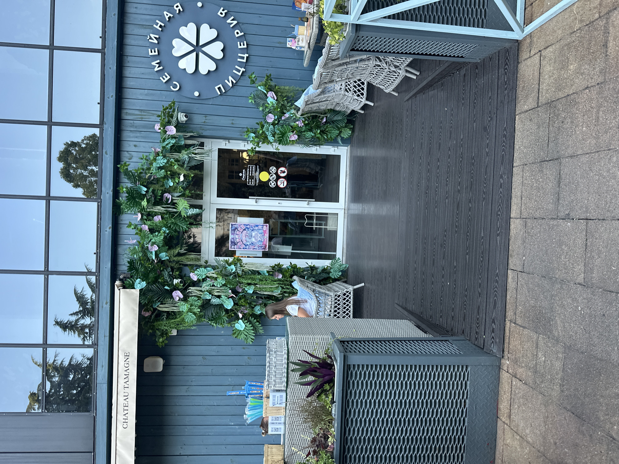 This media file captures an outdoor scene in front of a building with blue wooden siding. A large glass door with a white frame is the central element, decorated with abundant green foliage and some pink and light blue artificial flowers. To the left of the door, a large window with a grid pattern reflects trees. Above the door, a circular sign with white lettering and a flower-like design is visible.
In front of the door, a woman is seated in a white wicker chair, wearing a white top. She is looking down and appears relaxed. The area in front of the building is paved with dark wooden planks, and to the right, there is a brick wall. Two large, modern-looking planters with metal grates are also present. One planter contains green and purple plants.
On the left side of the building, a white awning is visible with the text "CHATEAU TAMAGNE" printed in black. Signs are attached to the glass door, one of which contains circular symbols, including one depicting a cigarette with a line through it, indicating a "no smoking" rule. Another sign appears to be a menu or informational poster. The overall impression is of a cafe or restaurant entrance, possibly in the afternoon based on the lighting and shadows. The setting appears to be Sochi, Russia, suggesting a potentially warm and pleasant day.