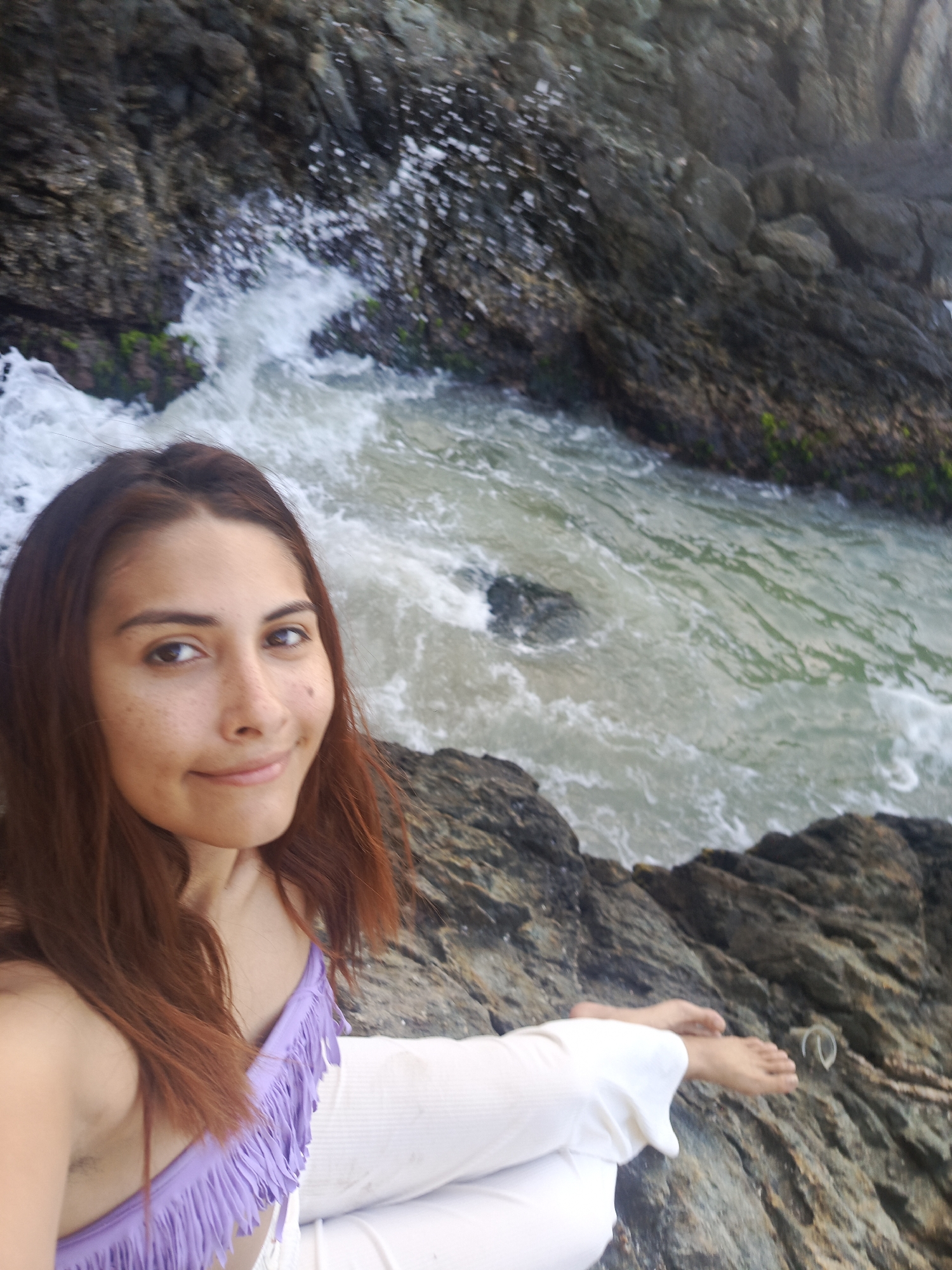 A woman with long, auburn hair and freckled skin smiles at the camera while sitting on a rocky shore. She is wearing a purple, fringed bikini top and white pants. The ocean is visible behind her, with waves crashing against the rocks. The sun is shining, casting a bright light on the scene. The overall impression is one of natural beauty and relaxation.
