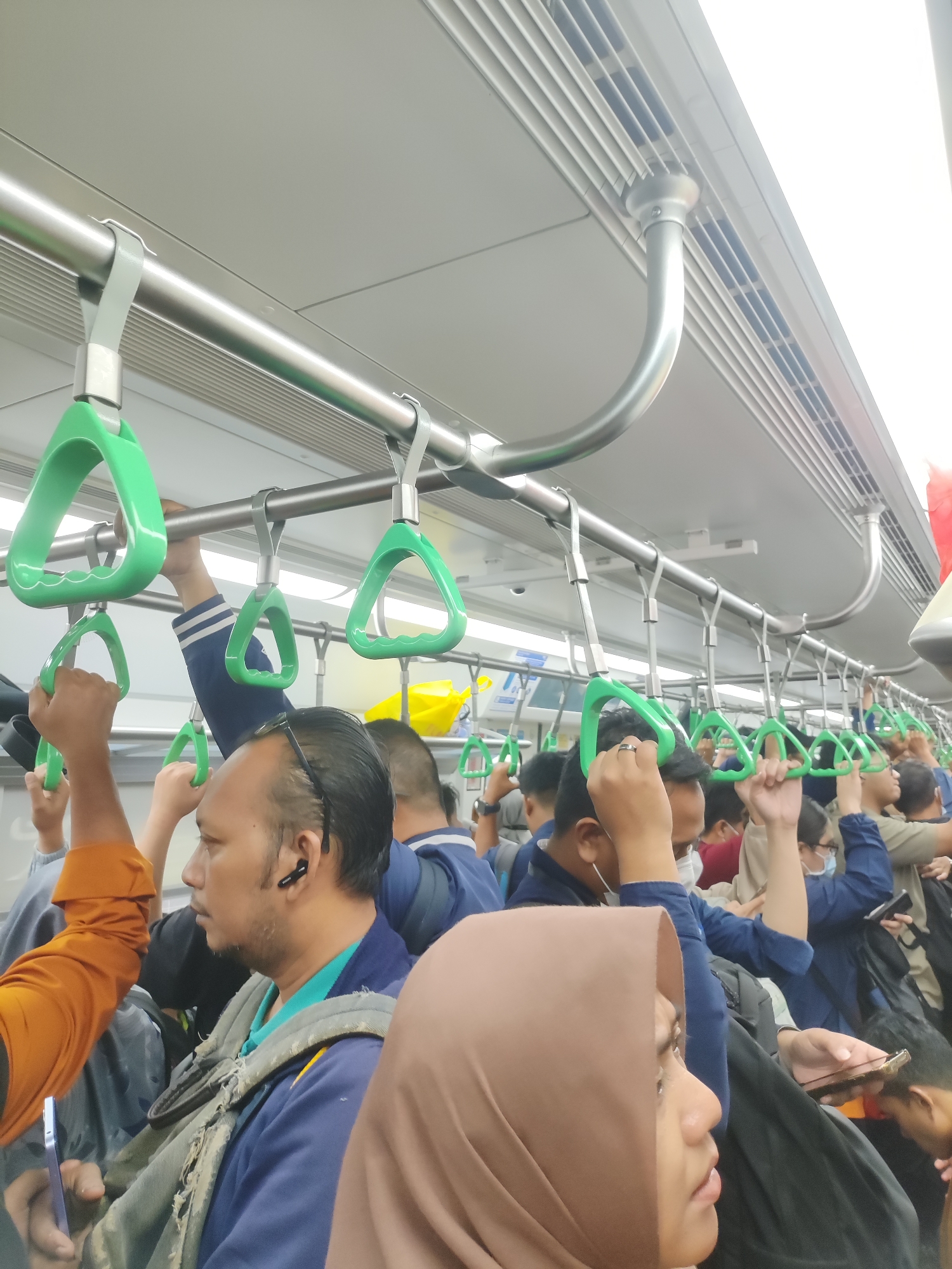 The image depicts the interior of a crowded commuter train, likely a KRL Commuter Line, with passengers holding onto green handgrips suspended from the ceiling. The scene is brightly lit, suggesting it is daytime, and the environment is clean and modern, typical of public transportation in Indonesia. Several individuals are visible, mostly facing forward or to the side, with a mix of expressions, indicating a typical commute. One person in the foreground is wearing a blue jacket and a backpack, while another person to their right is wearing a light brown hijab. The overall impression is one of a busy and active urban commute. The location is specified as the city of Bekasi, Indonesia, implying that this scene is representative of daily life in that region's public transit system.