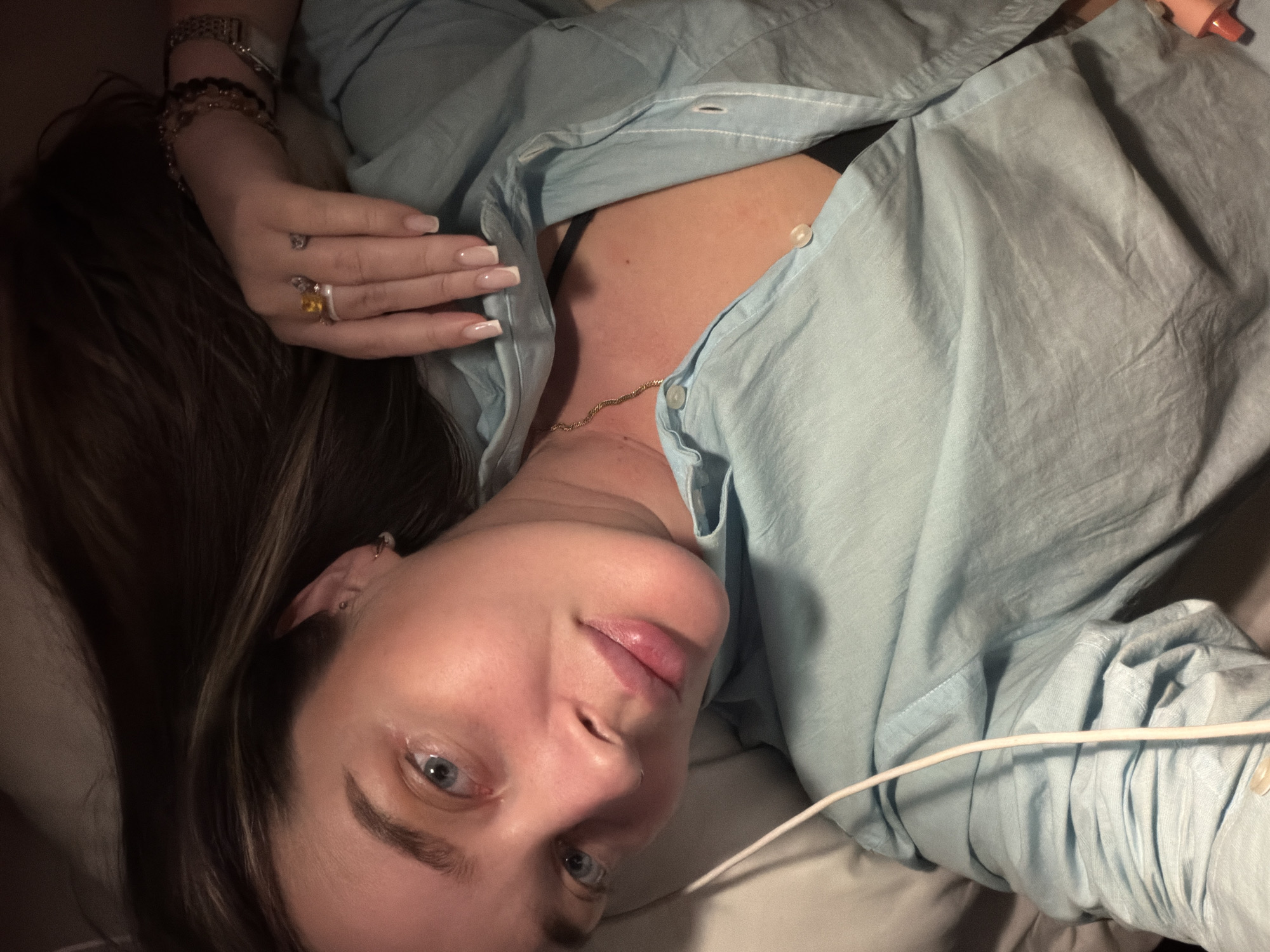 A woman with fair skin and blue eyes is lying down, wearing a light blue button-down shirt and a gold necklace. She has brown hair with blonde highlights, multiple rings on her left hand, and bracelets on her left wrist. A white cord is visible near the lower right corner. The location is not discernible as Moscow, Russia.
