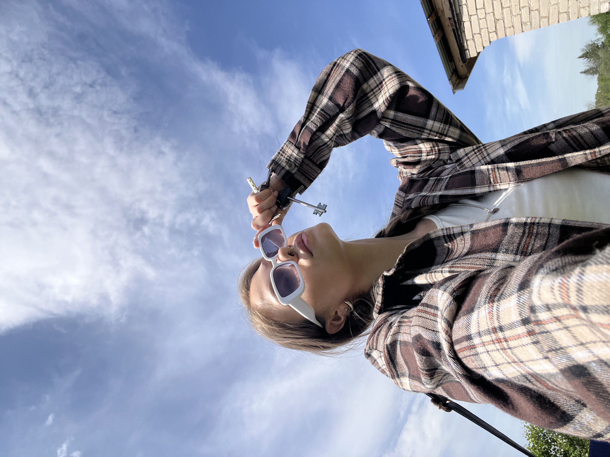 A low-angle, rotated photograph shows a young person against a blue sky with white clouds. The person is wearing white sunglasses and a brown and white plaid shirt over a white tank top. They hold a set of keys up near their forehead. At the top right of the image is a brick building and some trees. The location is unspecified.