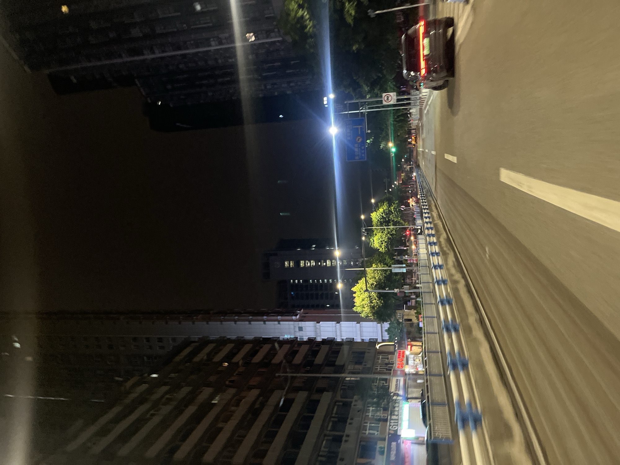 The image depicts a street scene at night in Jiulongpo District, China. Tall buildings line the left side of the road, with illuminated windows visible. A highway overpass with blue barriers runs along the left edge of the frame. The road ahead has multiple lanes, with white markings and some traffic. A dark-colored SUV is traveling in the right lane, with its taillights illuminated. Streetlights and traffic lights are visible, as are some trees and signs along the roadside. The overall impression is of a busy urban environment at night.