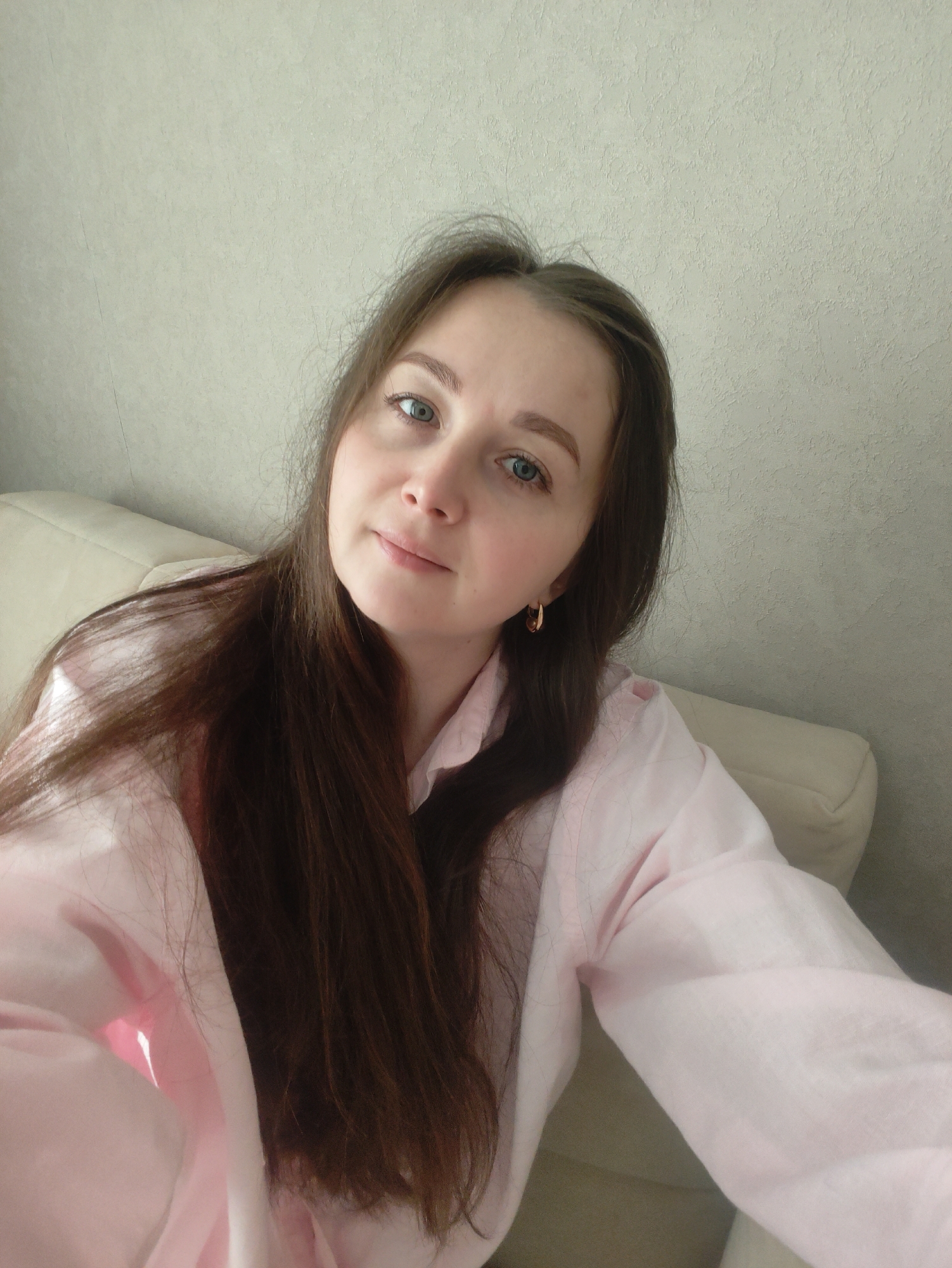 A young woman is captured in a close-up, eye-level selfie or portrait. She has long, straight, dark brown hair that cascades over her shoulders and chest, and striking blue eyes. Her eyebrows are light, and she wears a subtle, gentle smile, looking directly into the camera. She is wearing a light pink, possibly linen or cotton, long-sleeved shirt, and a gold-colored hoop earring is visible in her right ear.
She appears to be seated or reclining on a light beige or off-white sofa or armchair, which is partially visible on either side of her. The background features a light-colored wall with a subtle textured finish, suggesting an indoor setting, likely a home environment. The lighting is soft and natural, indicative of daytime. The overall mood is calm and pleasant. There is no visible text in the image.