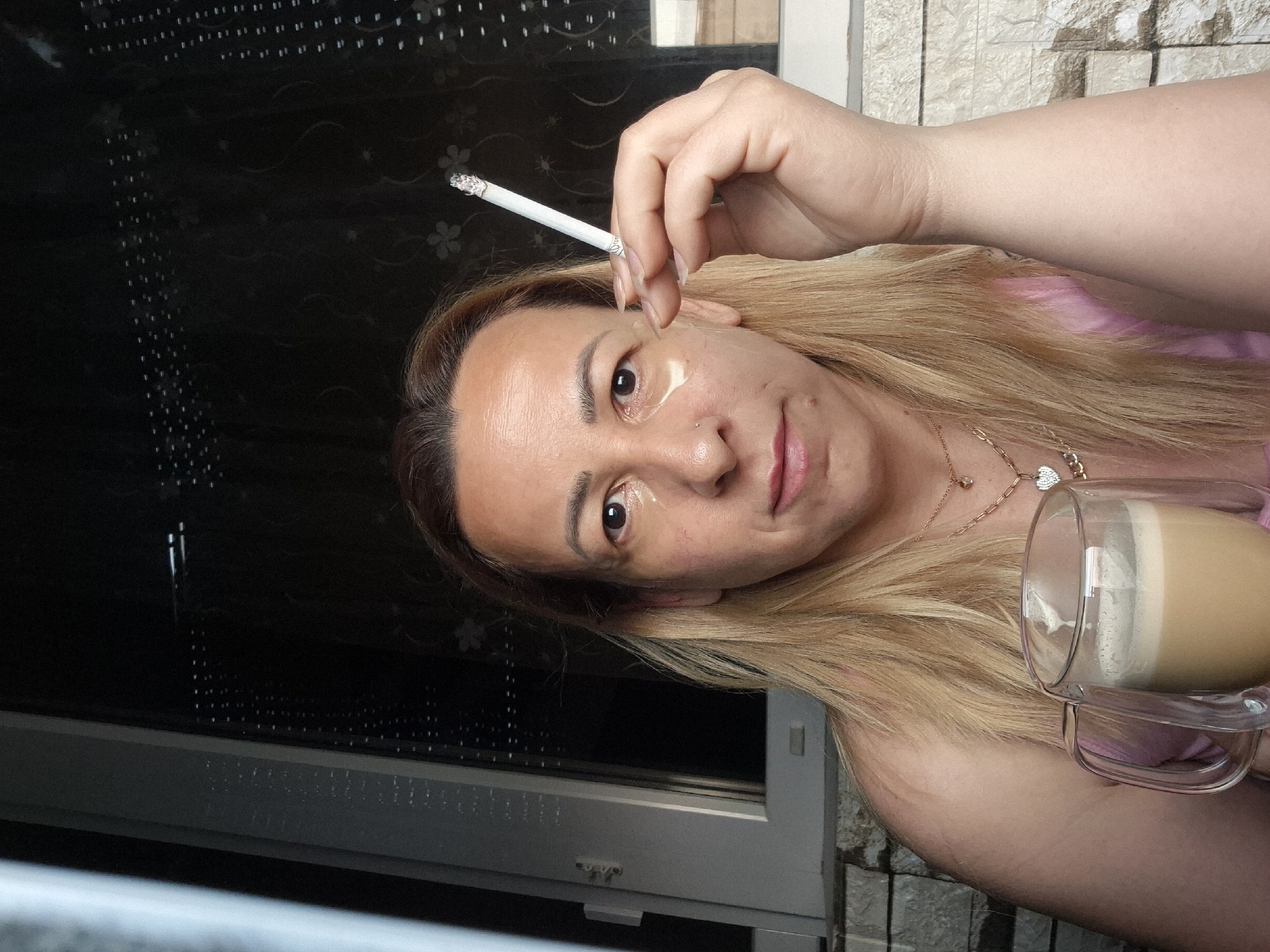 A medium shot captures a woman with blonde hair, wearing a pink top and gold necklaces. She holds a lit cigarette in her right hand, bringing it towards her face, and has eye patches under her eyes. To her left, she holds a clear glass mug filled with a frothy, light brown beverage. The background features a window with a patterned blind and a wall of textured stone blocks. The scene is outdoors, possibly on a balcony or patio.
