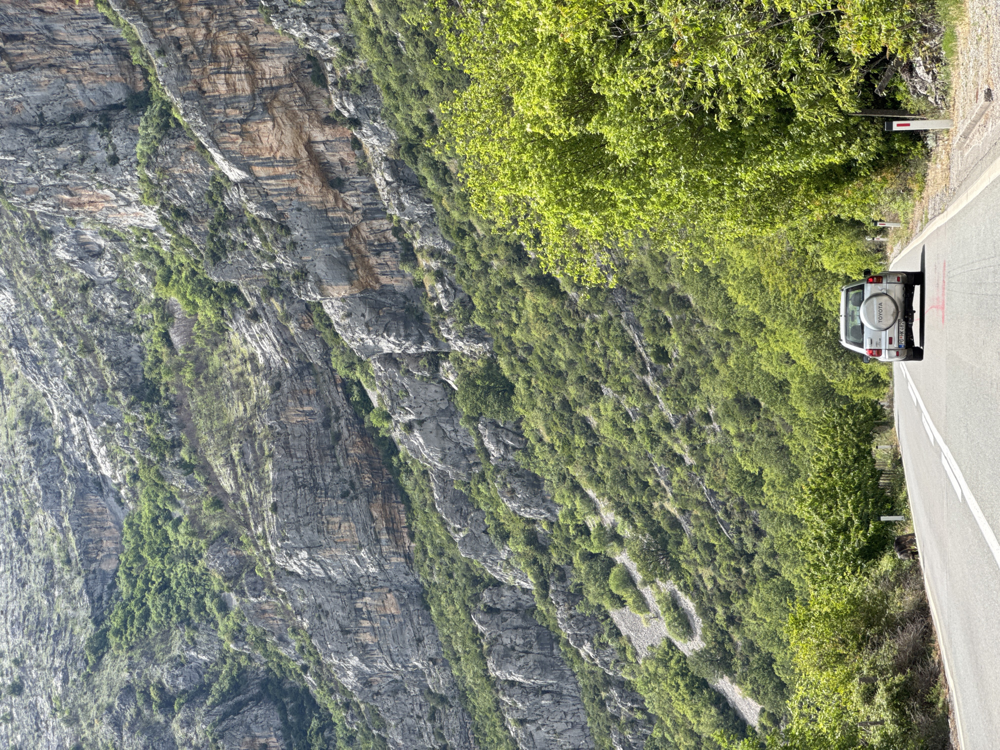 The image shows a silver Toyota Land Cruiser parked on a gray asphalt road alongside a steep, rocky mountainside with sparse green vegetation. The vehicle's license plate reads "PG-HK 482." A white line divides the road. The location is Donje Mrke, Montenegro, as evidenced by the license plate. The road is situated along a drop-off. A road sign is visible near the edge.