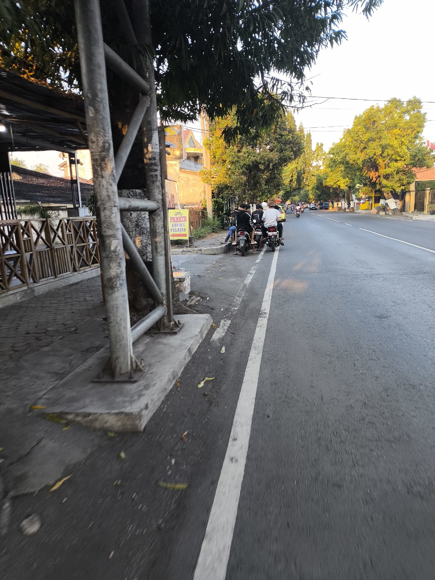 The image depicts a street scene in Citrodiwangsan, Indonesia, during daylight hours with the sun casting long shadows, suggesting it's either early morning or late afternoon. In the foreground, a concrete sidewalk runs alongside a paved road marked with a white line. A large metal pole, likely part of a shelter or overhead structure, stands prominently on the sidewalk.
To the left of the road, there is a wooden fence with a crisscross pattern, behind which appears to be an outdoor seating area or a cafe, with a dark-colored roof structure. A yellow sign with red text is visible on the sidewalk near the metal pole, indicating "PARKIR KHUSUS PELAJAR" which translates to "STUDENT PARKING ONLY," along with an arrow pointing to the right.
Several motorbikes are parked and occupied by people on the side of the road. Three individuals are seated on one motorbike, and two more on another. They appear to be dressed in casual attire, with some wearing white shirts. Further down the road, more motorbikes and some cars are visible, suggesting moderate traffic. Lush green trees line the right side of the road, some illuminated by the sunlight, creating a vibrant contrast with the asphalt and buildings. The buildings on the right appear to be low-rise structures with orange-tiled roofs. The overall impression is a typical street scene in a populated area of Indonesia, likely near a school or place of business.
