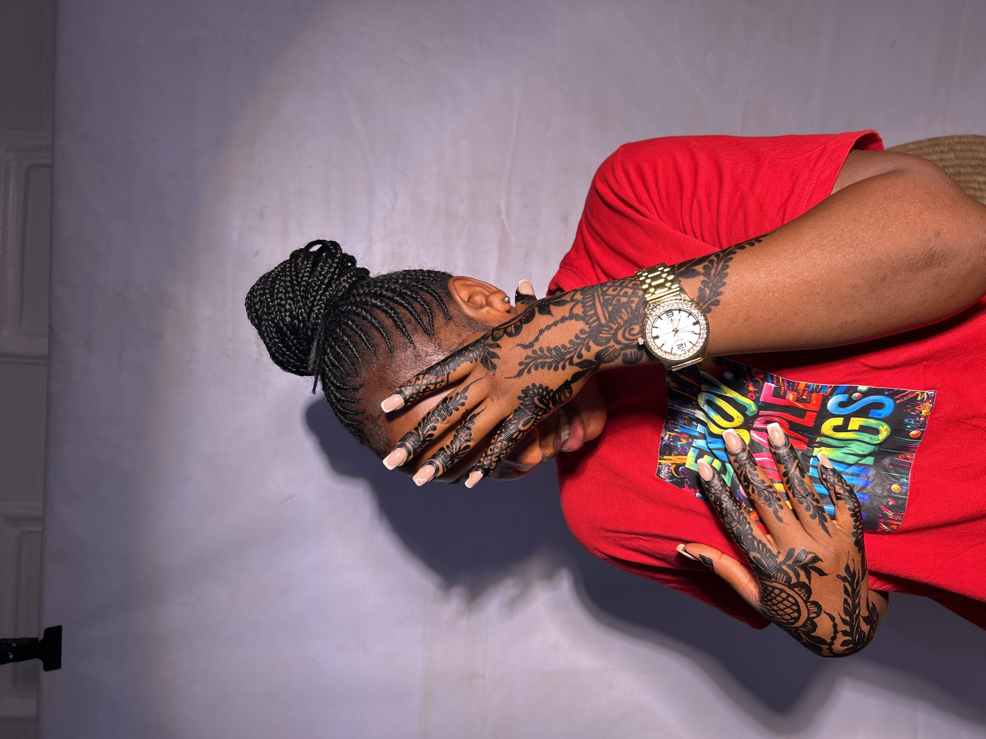 A person is depicted in a studio setting against a light gray background. The individual is wearing a red T-shirt with a colorful graphic design on the front. The person's hands, adorned with intricate henna designs, are raised and partially covering their face. The hair is styled in braids, gathered into a bun. A gold wristwatch is visible on one wrist. The location in the city of Jos, Nigeria, cannot be verified based on the given image.