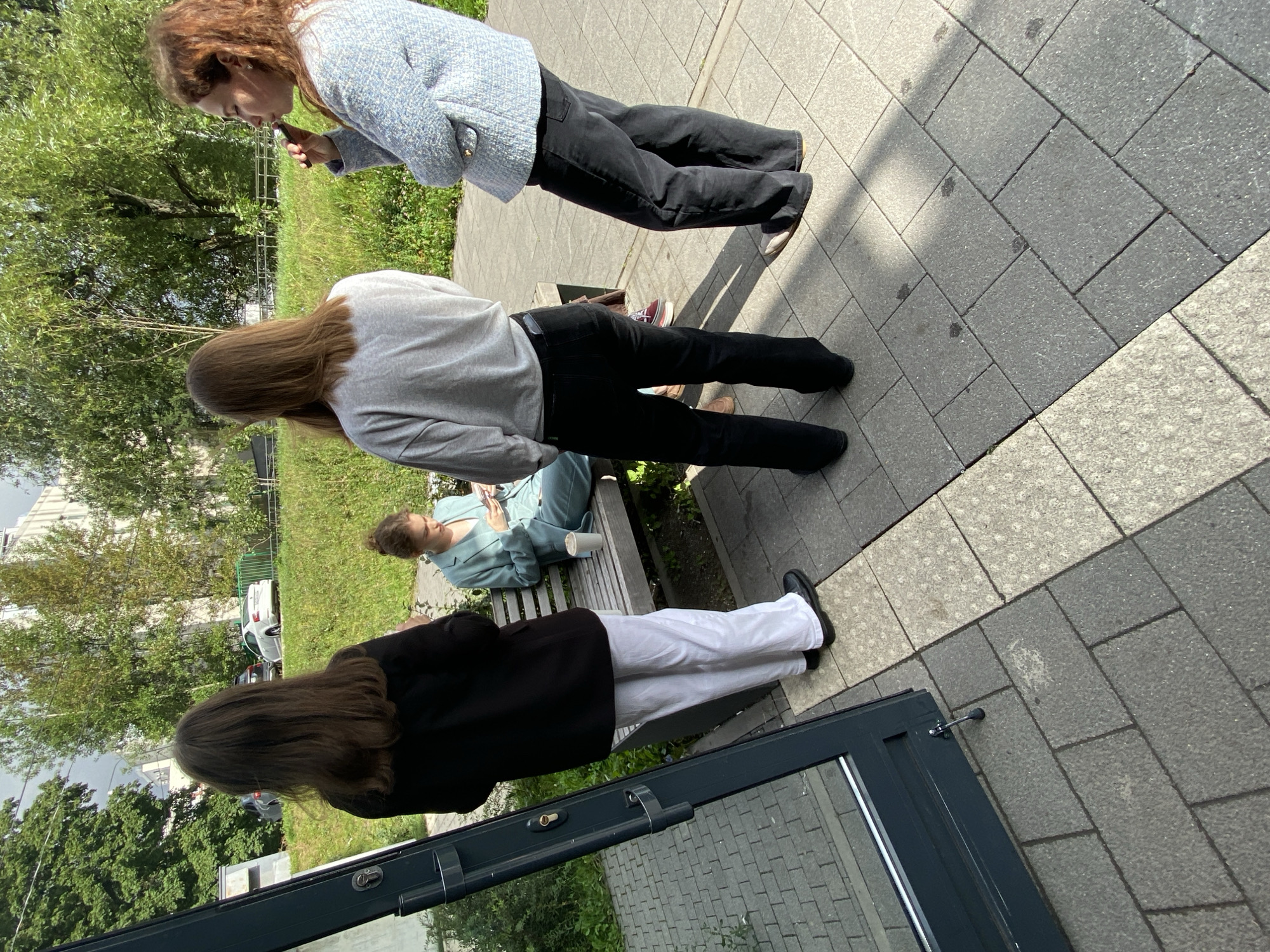 The image depicts an outdoor scene with four individuals and architectural elements. In the foreground, to the left, is a dark gray door with a glass panel, revealing a reflective surface. The ground is paved with gray and light-colored square tiles, forming a pathway.
Further into the scene, a woman with long brown hair, wearing a black blazer and white pants, stands with her back to the camera. To her right, a woman with brown hair, wearing a light gray sweatshirt and black pants, is also viewed from behind. In the center, a woman in a light teal pantsuit sits on a wooden bench, looking at her phone. Beside her on the bench is a white paper cup. To the far right, a woman with reddish-brown curly hair, wearing a light blue textured jacket and black pants, is seen from the side, holding a dark cylindrical object to her mouth, possibly a vape pen.
The background features a grassy slope with trees and some buildings. The overall impression is a casual outdoor gathering, possibly near a building entrance or a public waiting area. The lighting suggests it is daytime, with shadows cast on the pavement. The image does not provide specific clues to pinpoint the exact location within Moscow, Russia.