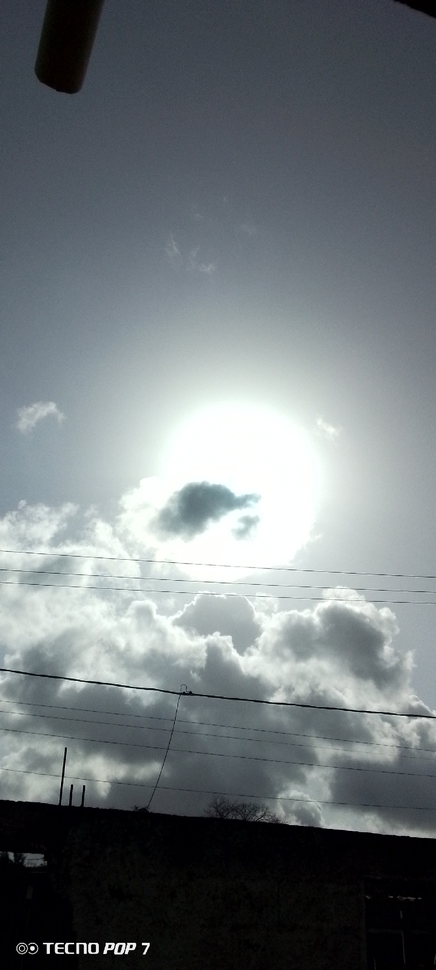 The image depicts a daytime sky with bright sunshine partially obscured by clouds. There are multiple layers of clouds, with darker, heavier clouds in the lower portion of the frame. Overhead power lines traverse the middle ground. In the foreground, there is a dark structure, possibly a building or a wall, with a small tree partially visible. The location information, "TECNO POP 7," likely indicates the device used to take the photo and does not relate to geographic information. There are no people visible in the image. The setting is a cloudy day, and the environment is urban with power lines and buildings present. Altagracia, Venezuela, is not visually discernable in the image alone, but could be the place where the photo was taken.