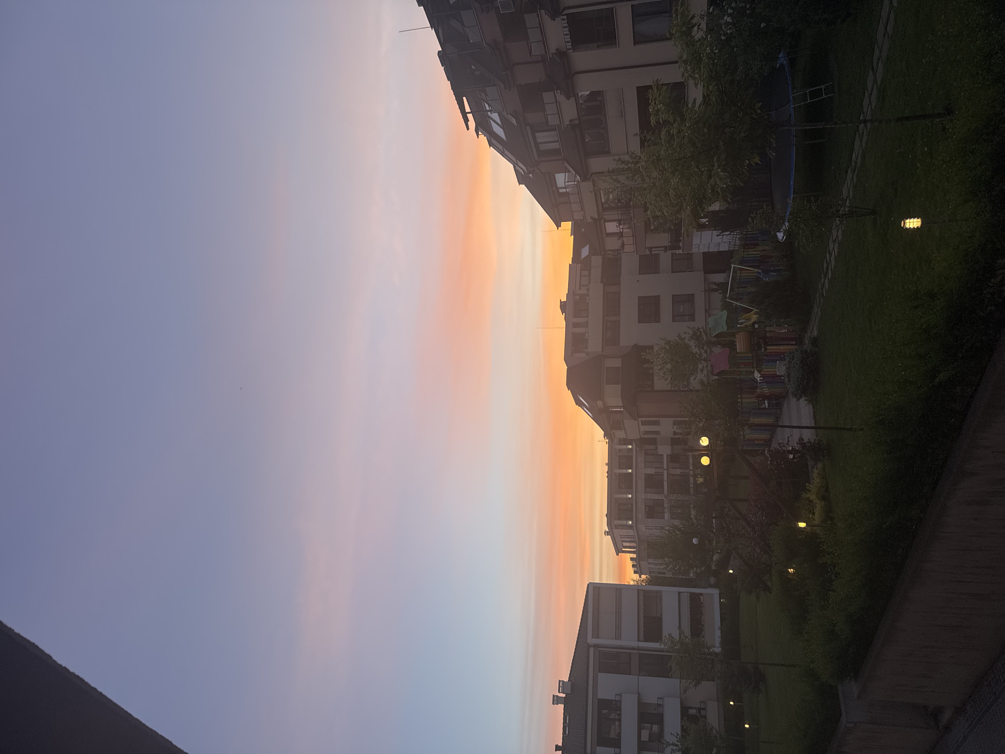The image shows a view of apartment buildings in a neighborhood in Sofia, Bulgaria, at dusk or dawn. The sky is a gradient of light blue to orange, indicating either sunrise or sunset. The buildings are of modern construction, with multiple stories and balconies. A small park or green area is visible between the buildings, with trees, lamps, and what appears to be playground equipment. The lighting suggests it's early morning or late evening, with some artificial lights turned on. No people are immediately visible in the shot.