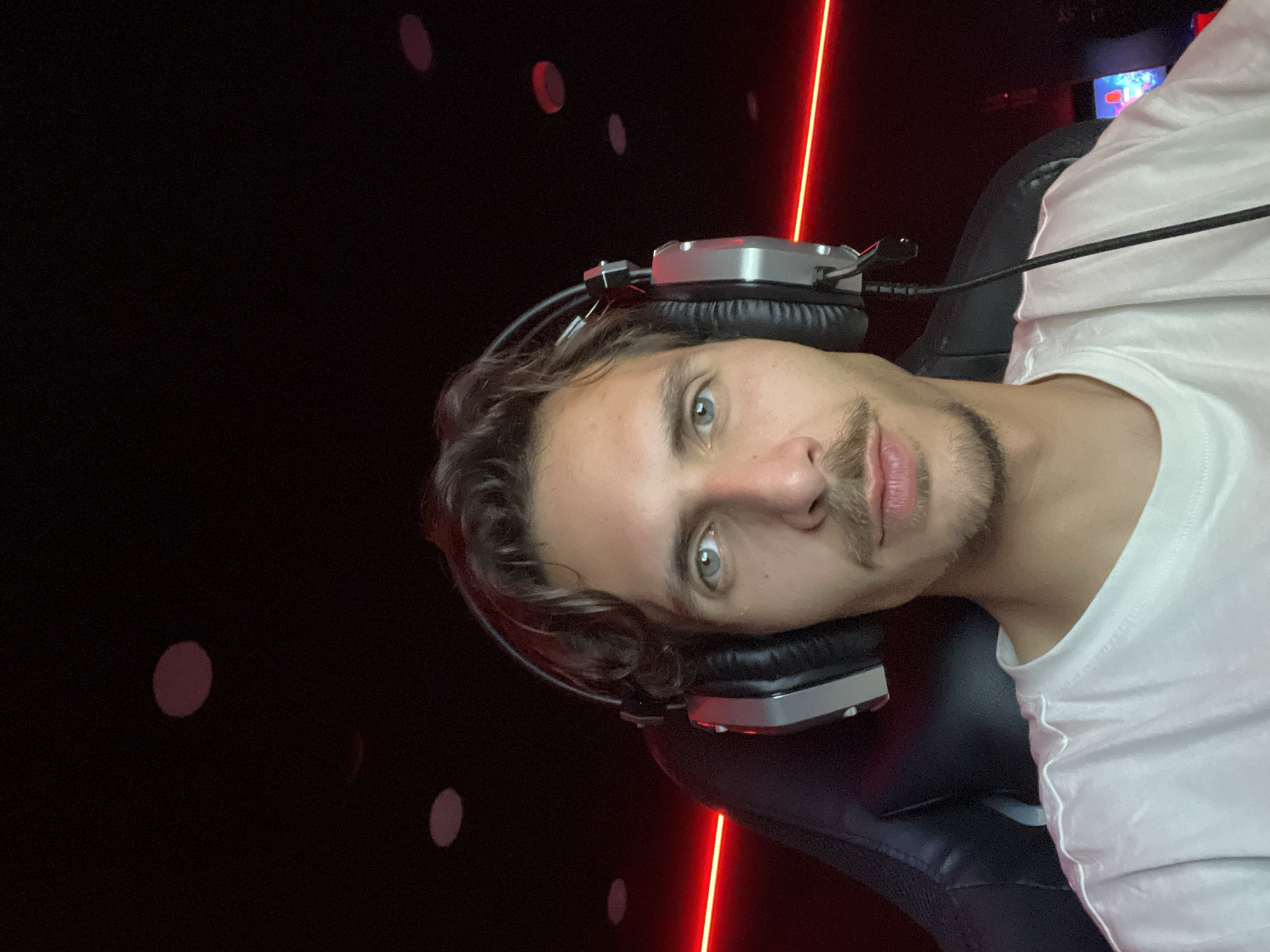 A person is seated in a black chair, wearing a white shirt and headphones. The background is dark with several indistinct, reddish-pink circular shapes and a prominent vertical red light streak. The individual has light eyes and a short, neatly groomed mustache and beard. The photograph appears to be taken indoors, but the exact location within Budva, Montenegro cannot be determined from the image.