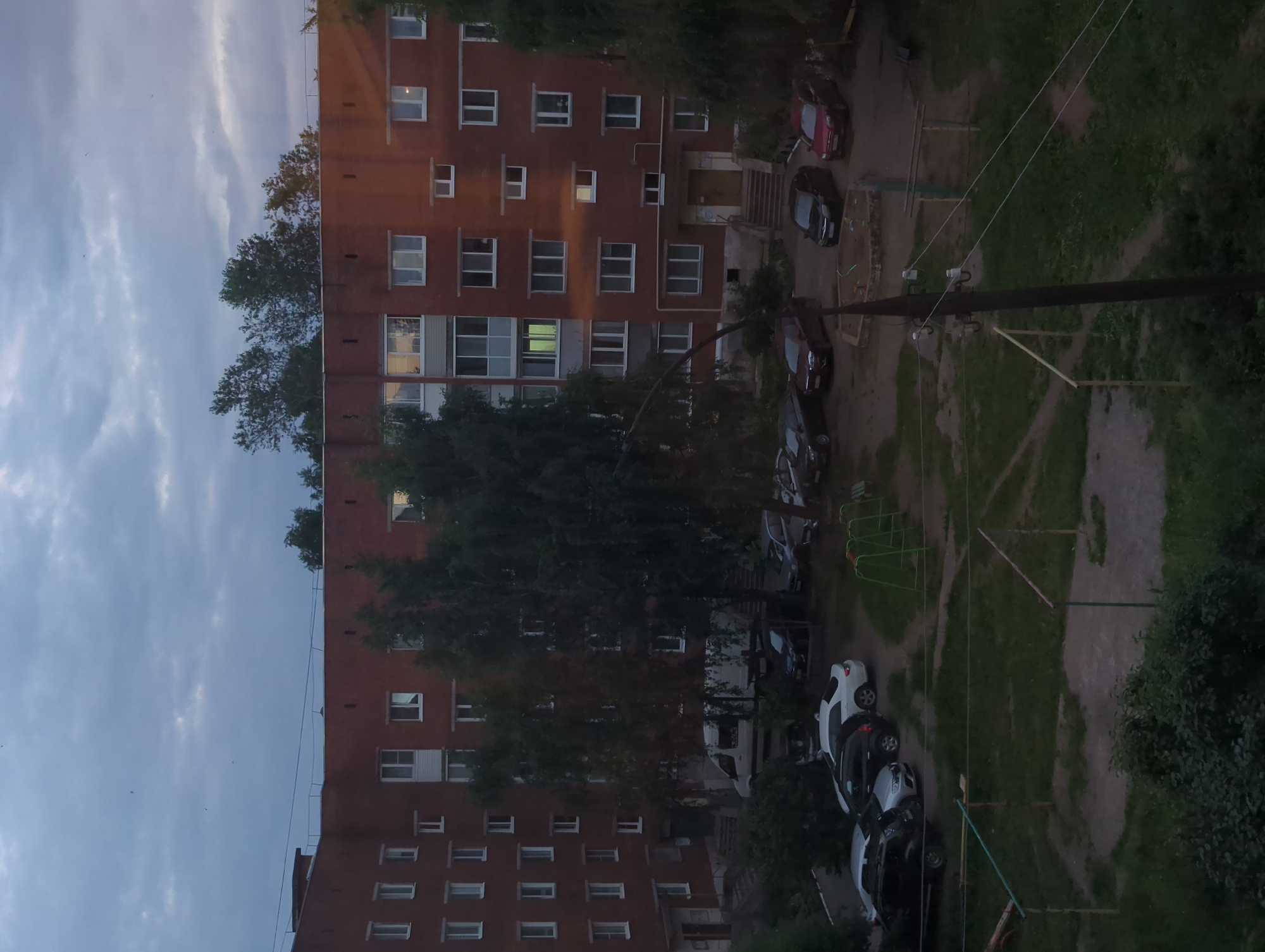 The image shows a residential courtyard in Votkinsk, Russia, during dusk. A multi-story brick apartment building dominates the scene, with numerous windows visible. Several cars are parked in the courtyard, including a white sedan, a black SUV, and a red car. There is a children's playground with swings and a sandpit. Trees and bushes are scattered throughout the courtyard, and some power lines are visible. The sky is overcast, suggesting an approaching rain or evening. No people are visible in the image.