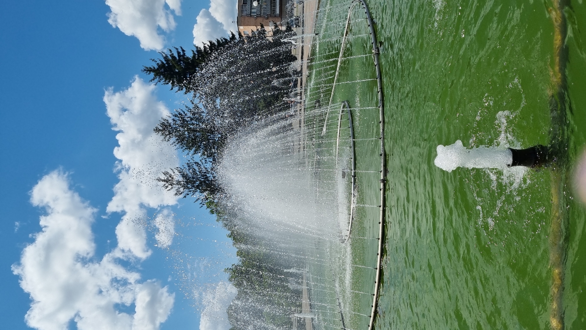 The image depicts a public fountain in the city of Smila, Ukraine, on a clear day with scattered white clouds against a blue sky. The fountain is active, with water jets creating a large spray in the center and smaller jets around the perimeter. The water in the fountain basin is a vibrant green. In the background, there are several tall evergreen trees and a multi-story building. A paved area, likely a promenade or square, is visible behind the fountain, with a few indistinct figures of people walking. The image is captured from a low angle, focusing on the fountain and its spray.