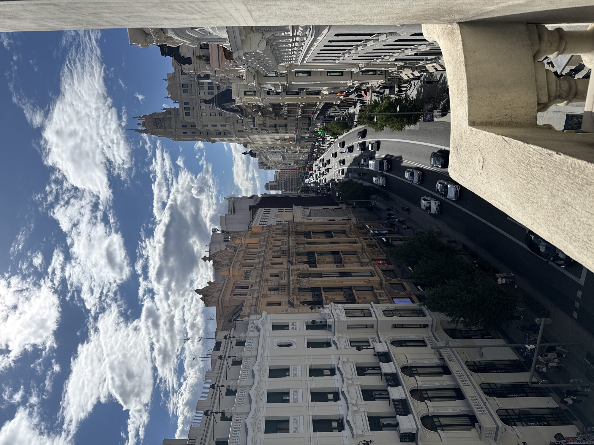 The image depicts a view of Gran Vía, a prominent street in Madrid, Spain, taken from an elevated perspective, possibly from a balcony or a high-rise building. The street is busy with vehicular traffic. Several cars and scooters are visible moving along the road. Buildings with ornate façades line both sides of the street. The architectural style is predominantly historical, with detailed decorations and elaborate window designs. The sky is partly cloudy, with patches of blue visible among the white clouds. A stone railing or architectural detail is visible in the foreground, indicating the viewpoint. People can be seen walking on the sidewalks on either side of the street.