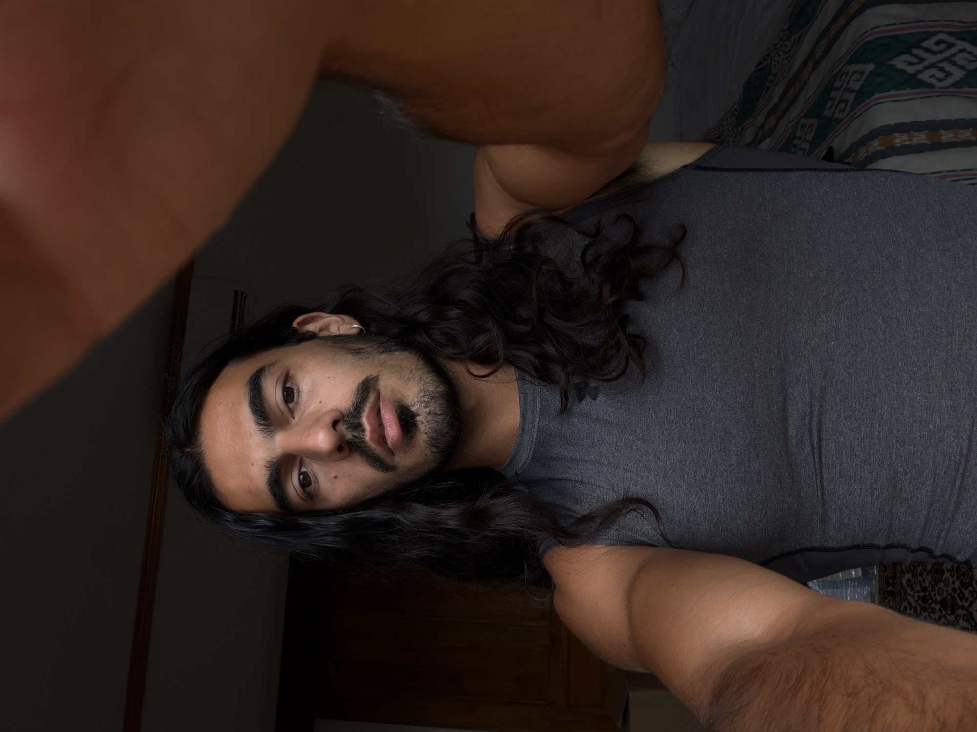 A man is lying on a bed, taking a selfie. He has long, dark, curly hair, a beard, and an earring in his left ear. He is wearing a gray t-shirt. The bed has a patterned blanket on it. The location may be in North Kuta, Indonesia.