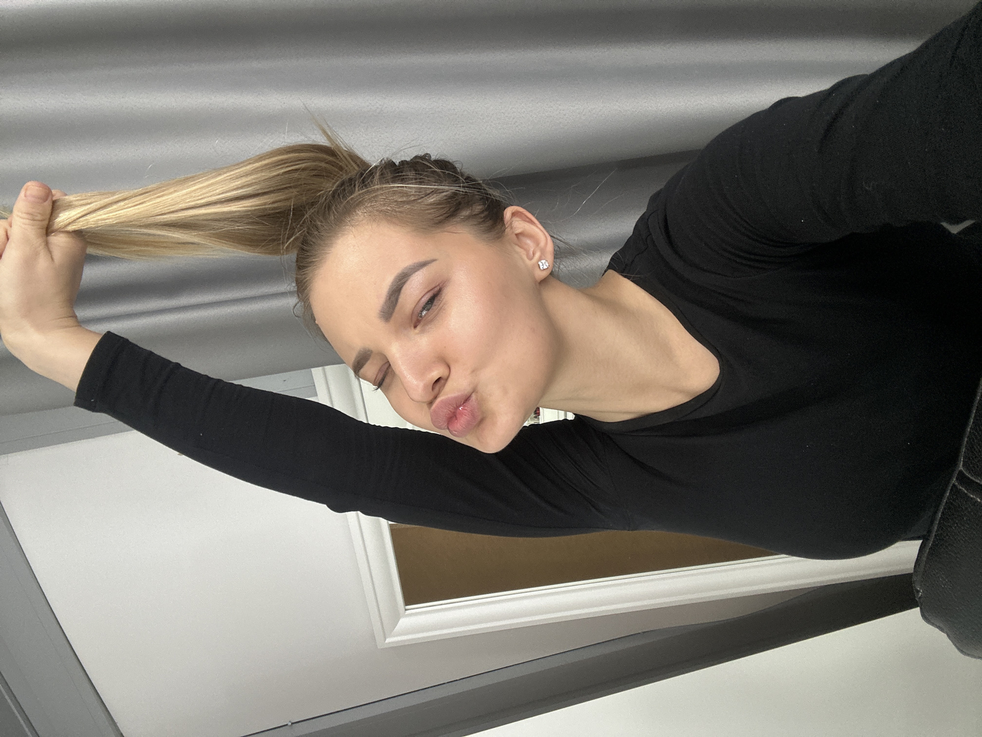 A young woman with light skin and blonde hair pulled into a high ponytail is holding the end of her ponytail in her left hand. She is wearing a black long-sleeved shirt and a small square earring in her left ear. She is making a kissing face with her lips puckered. The background includes a gray textured surface at the top and a white door frame with a brown interior visible behind it. The location is not discernible from the provided image details; hence, it cannot be confirmed if this image was taken in Budva, Montenegro.