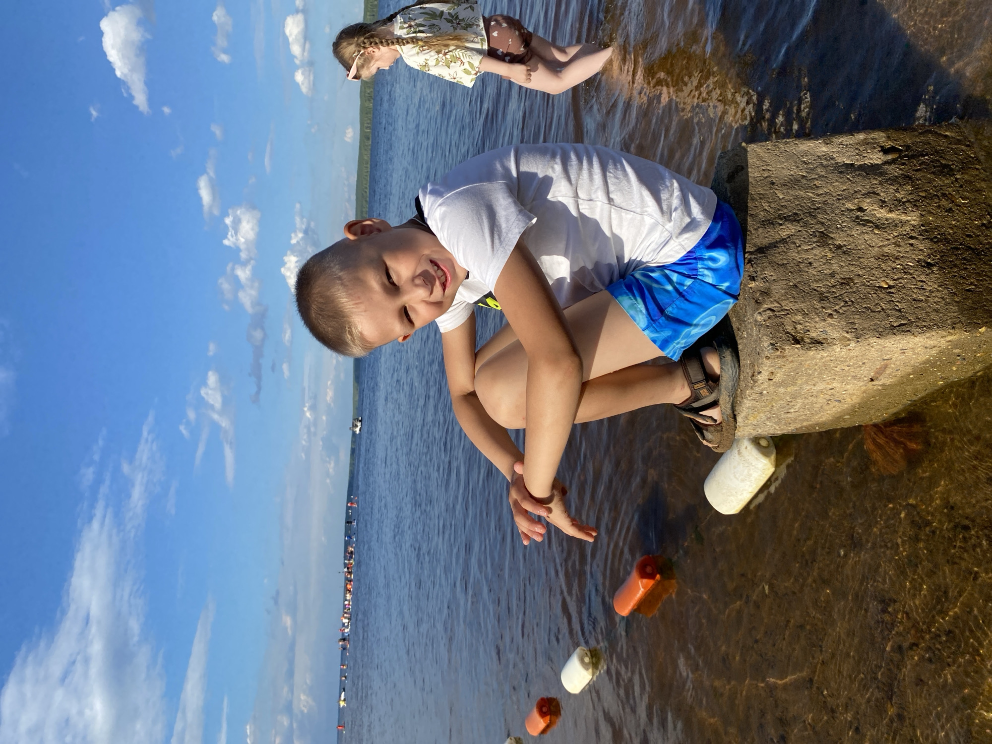 The image captures a bright, sunny day at a body of water, likely a river or lake, near the city of Chaikovsky, Russia. In the foreground, a young boy with short, light-colored hair and a bright smile is seated on a concrete block at the water's edge. He is wearing a white t-shirt and blue shorts with a swirling pattern. His hands are resting on his knees, and he is looking directly at the camera. To his right, in the background, a young girl with braided hair is standing in the shallow water, facing away from the viewer. She is wearing a patterned top and brown bottoms. Several orange and white buoys are visible floating in the water near the shore, marking a boundary or a course. Further out on the water, a distant group of people on various watercraft, possibly kayaks or paddleboards, are visible, along with a small boat. The sky is a vivid blue with scattered white clouds, and a treeline can be seen in the far distance. The water is calm and reflects the sunlight.