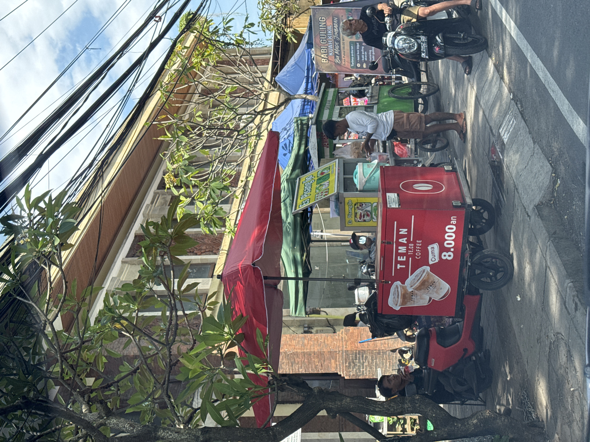 The scene shows a bustling street in Kuta Selatan, Indonesia, viewed from a vehicle. A red mobile coffee cart, branded "TEMAN TE.CO COFFEE," is positioned on the sidewalk. A vendor in a white shirt prepares a drink. Next to the coffee cart is an "Es Pisang Ijo" food stall. Behind it, another food stall selling "BABI GULING KUAH KIKIL" is visible. An elderly man on a motorcycle, possibly a customer, is parked nearby. Power lines and buildings dominate the background. The street is marked with a white line. A tree with foliage is present in the foreground.