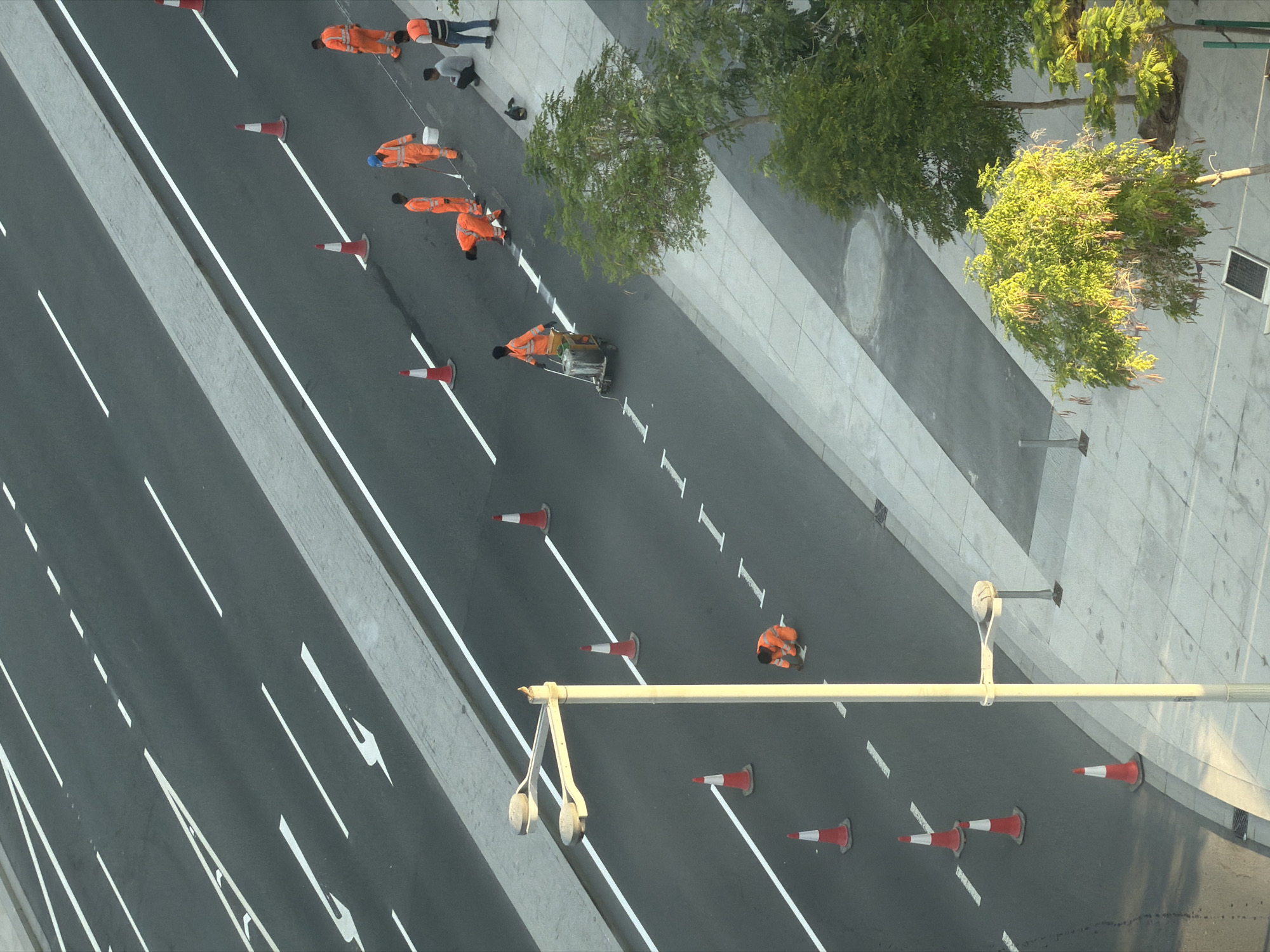 An overhead, eye-level shot captures a team of road workers in orange jumpsuits performing line painting on a multi-lane asphalt road in an urban environment. The road features directional arrows and white dashed and solid lines indicating lanes. Orange traffic cones are strategically placed to demarcate the work area. One worker operates a line-painting machine, while others appear to be applying paint manually with brushes, stooping down to touch up markings. Several workers are gathered, some observing the process, while two individuals in casual attire sit on the sidewalk adjacent to the road. A lamppost stands prominently on the left side of the road. Trees with green foliage are visible on the right side of the frame, bordering a paved walkway. The scene suggests ongoing road maintenance or construction work.