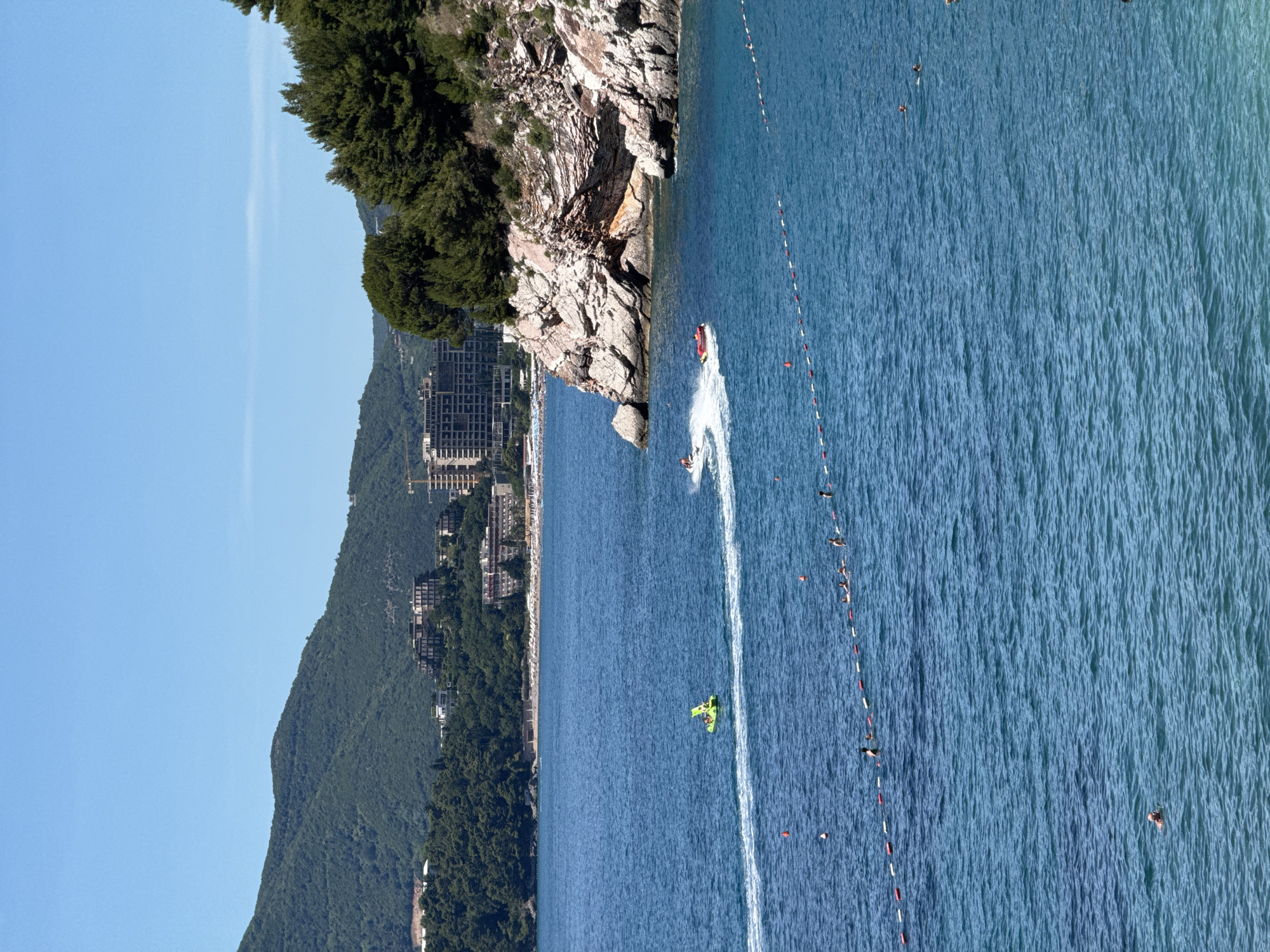 The image depicts a coastal scene in Kamenovo, Montenegro, on a clear, sunny day. The turquoise water of the Adriatic Sea dominates the foreground, with small waves and ripples. A yellow inflatable watercraft, possibly a jet ski or a towed inflatable, is being ridden by at least one person, leaving a white wake. Further in the mid-ground, another person on a jet ski creates a more substantial wake as they move towards the right side of the frame, past a red inflatable object. A rope with alternating red and white buoys delineates a swimming area in the water. Several people are visible swimming and wading within this roped-off section. In the background, a rocky, tree-covered cliff face juts out into the sea on the right. Behind the cliff and extending across the horizon are lush green hills and mountains, dotted with buildings and hotels, indicative of a resort town. The sky is a clear, bright blue.