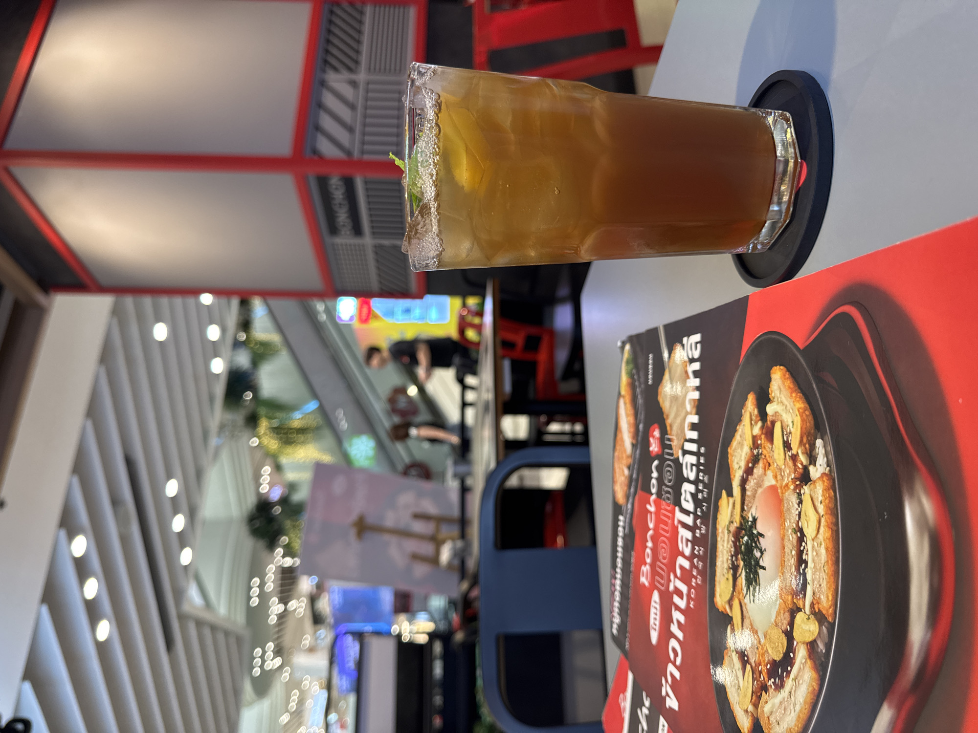 The image shows a close-up of a tall glass of iced tea with mint and lemon slices, placed on a black coaster on a grey table. In the foreground, a menu is partially visible, displaying images of Korean food, specifically rice bowls topped with meat and a fried egg, and the Bonchon logo. The background depicts a bright, modern indoor setting, likely a restaurant or food court in a shopping mall in Bangkok, Thailand, with escalators, seating areas, and overhead lighting. The overall atmosphere suggests a casual dining experience.