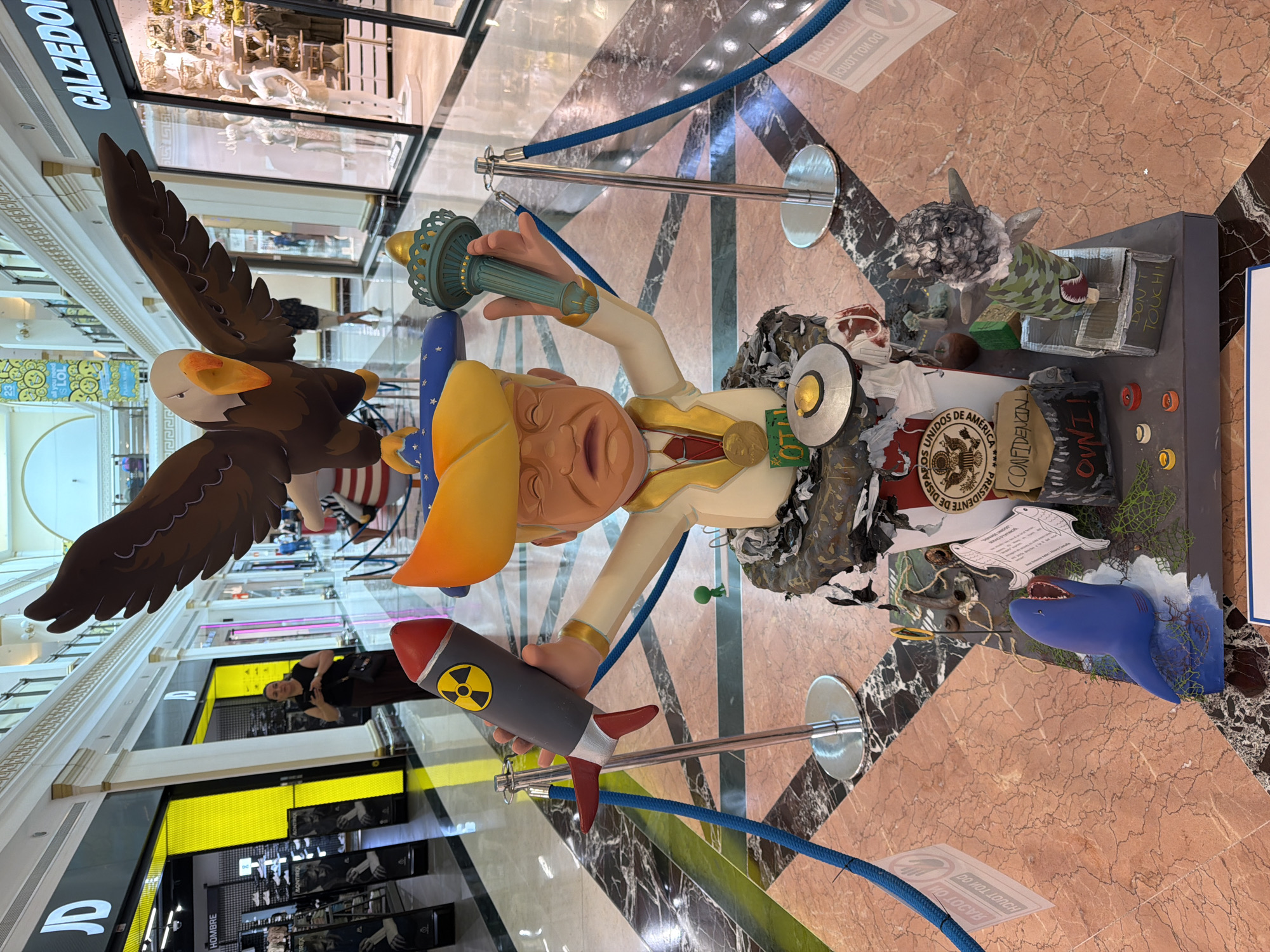 A large, elaborate, satirical sculpture is displayed in a shopping mall. The sculpture features a caricature of a man with blonde hair wearing a golden blazer, a red tie, and a yellow hat adorned with stars. He is depicted with his eyes closed and mouth open in a distressed expression. In his right hand, he holds a replica of the Statue of Liberty's torch, and in his left hand, he holds a model of a rocket with a radioactive symbol.
The figure is positioned atop a base that appears to be a trash bin filled with debris, including medical masks and what seems to be a piece of meat. Various other objects are scattered around the base: a blue shark figure with its mouth open, a smaller shark figure with a camouflage pattern and missiles, a circular metallic object, and several small colored discs. Some of these items have text labels, such as "CONFIDENTI", "OWN IT", and "DON'T TOUCH!". The sculpture is roped off with stanchions and blue ropes.
In the background, the interior of a modern shopping mall is visible, with shops lining the walkway. The floor is a polished marble with a pinkish hue and dark veins. Several people are present in the mall, though none are directly interacting with the sculpture. The lighting is bright and commercial, consistent with a retail environment. The overall scene suggests a public display of art or protest, likely political in nature, given the imagery.