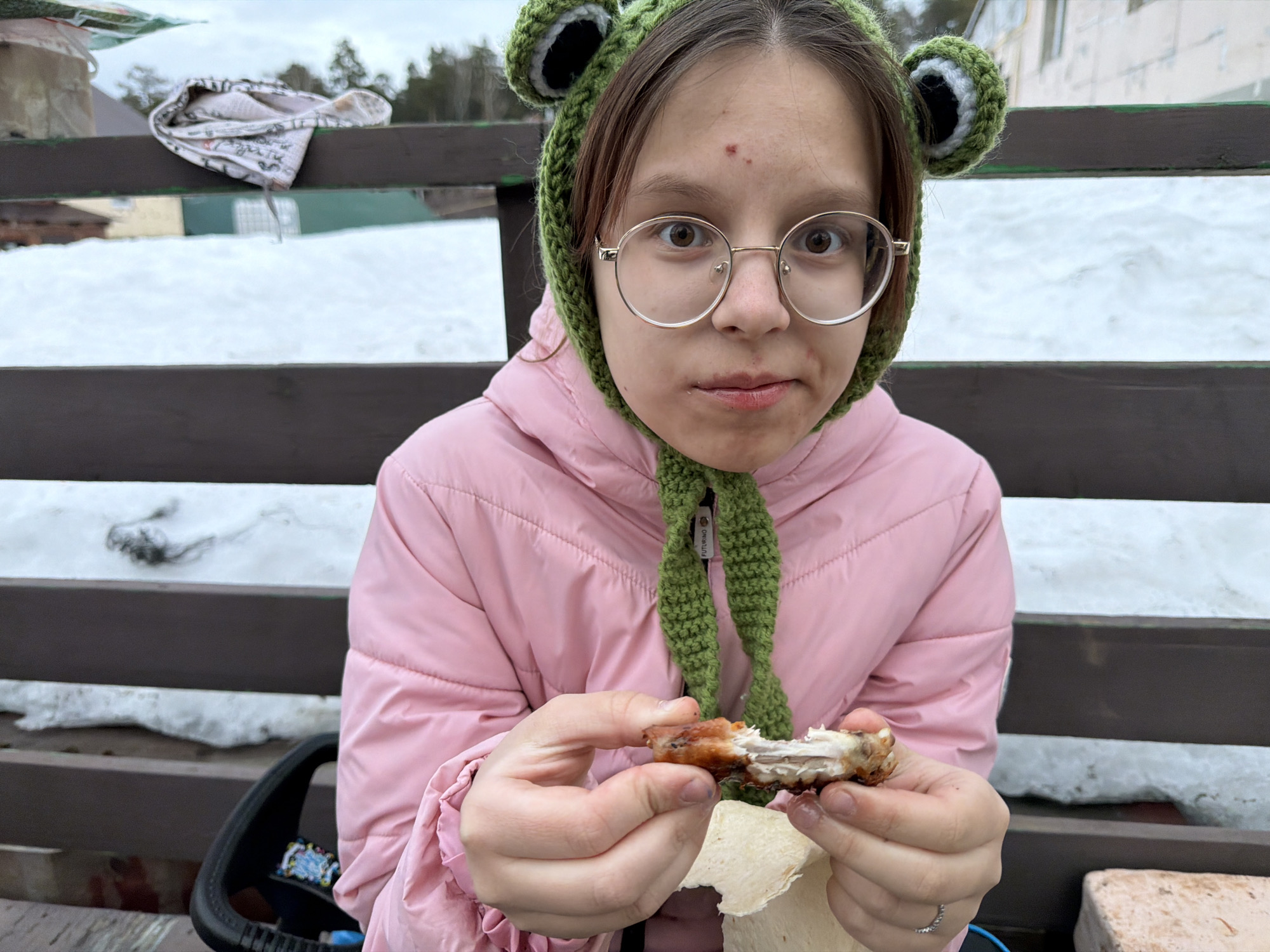 The image depicts a young female, approximately adolescent, situated outdoors in Chaikovsky, Russia, during what appears to be winter or early spring due to snow cover. She is positioned centrally, facing directly towards the viewer. She wears a light pink puffer jacket and a dark green crocheted hat with distinct frog-like ear elements featuring white and black centers, secured by ties under her chin. Round, gold-rimmed spectacles rest on her face. Two small, reddish blemishes are visible on her forehead, and her lips show a slight red discoloration. In her right hand, she holds a partially consumed piece of grilled poultry, exposing bone and white meat. Her left hand holds a folded piece of flatbread or tortilla. Her mouth is slightly open, suggesting she is in the process of eating. A silver-colored ring with small stones is on her left ring finger.
Behind the individual is a dark brown wooden railing or fence. Beyond this railing, the ground is covered extensively with white snow. In the far background, blurred coniferous trees are visible under an overcast, light grey sky. A light-colored building structure is faintly discernible behind the trees on the left side of the frame. A crumpled white cloth item with a faint pattern is draped over the wooden railing to the left. A black object, possibly a component of outdoor furniture, is visible in the lower left quadrant of the image. A light-colored block, possibly concrete, rests on the snow-covered ground in the lower right corner.