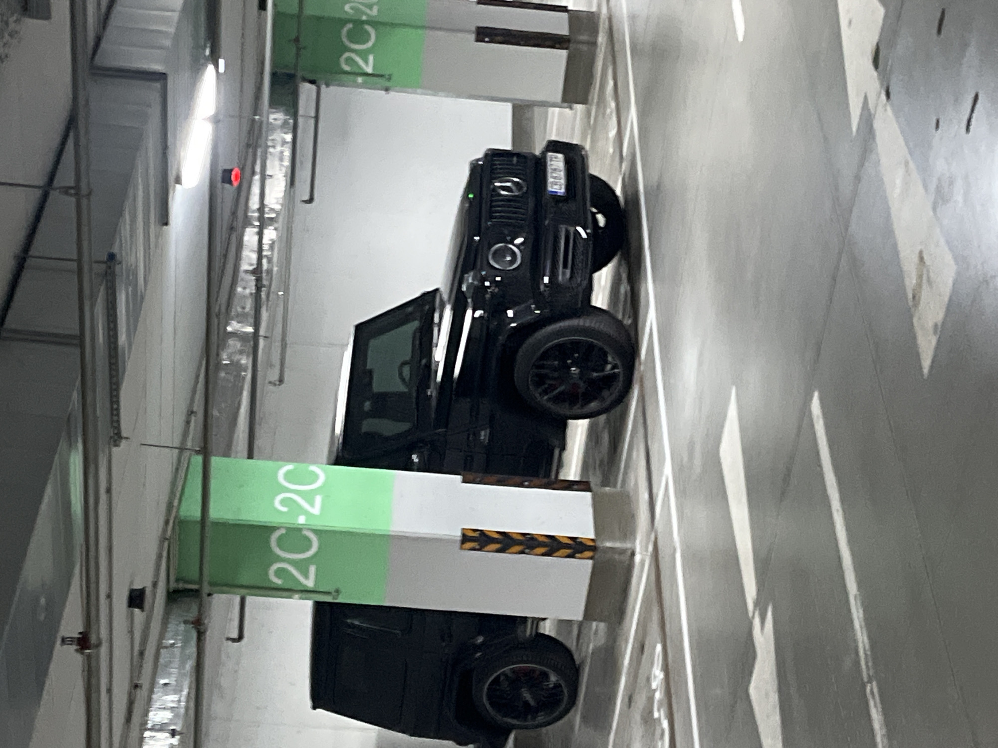 The image captures an indoor parking garage, likely an underground or multi-story facility, characterized by concrete structures and artificial lighting. The floor is a dark gray asphalt or concrete, marked with white painted lines defining parking spaces and directional arrows.
The primary subject is a black Mercedes-Benz G-Class SUV, parked in a designated space. The front of the vehicle is visible, featuring its iconic grille with the Mercedes-Benz emblem, circular headlights, and dark alloy wheels. The license plate, "C 8798 TP", is clearly displayed on the front bumper, indicating a Bulgarian registration from Sofia.
Two large rectangular pillars are visible. The one closer to the viewer has a prominent green section at its top with the white text "2C-2C," followed by a white section wrapped with yellow and black chevron-patterned hazard tape. A similar green-banded pillar, also marked "2C-2C," stands further in the background.
Overhead, various pipes, conduits, and ventilation ducts run along the ceiling. Artificial lights illuminate the area, with one bright light source and a smaller red indicator light visible on the wall. There are no people present, and the scene depicts a static moment with the car simply parked. The location context is Sofia, Bulgaria, as confirmed by the license plate.