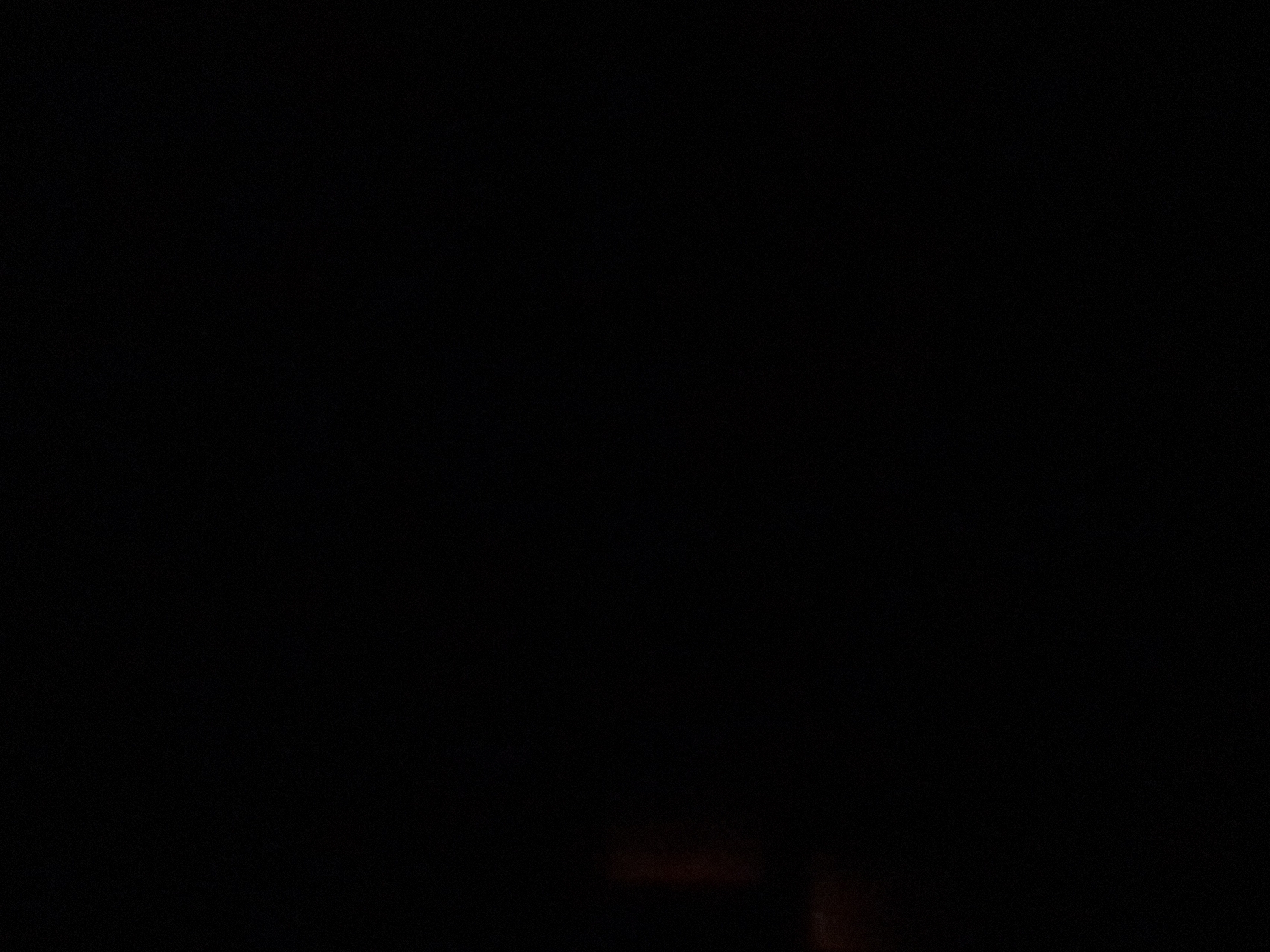 The image is extremely dark, with very little visible detail. In the upper right quadrant, there is a faint glow of reddish-orange light, suggesting a distant light source or reflection. The rest of the image is predominantly black, making it impossible to discern any specific objects, people, or environmental features. There is no discernible context related to the city of Orzhiv, Ukraine.