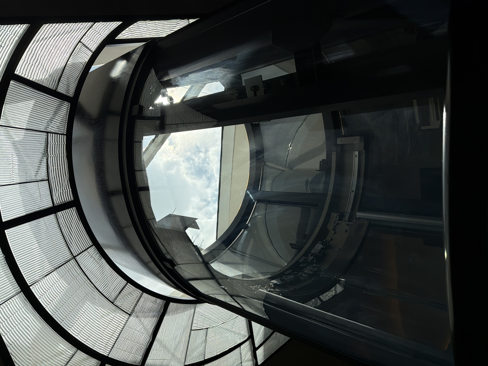 An upward, wide-angle, low-angle shot shows the interior of a cylindrical glass elevator shaft with a segmented glass ceiling. The elevator shaft is made of curved glass panels supported by dark metal framing. Through the glass panels, the sky with white clouds is visible. The ceiling is a series of ribbed glass panels set within a black metal frame. The scene is dimly lit, with light filtering in from the open ceiling and the glass enclosure. The image appears to be taken from inside the elevator car, looking upwards. The location is unknown.