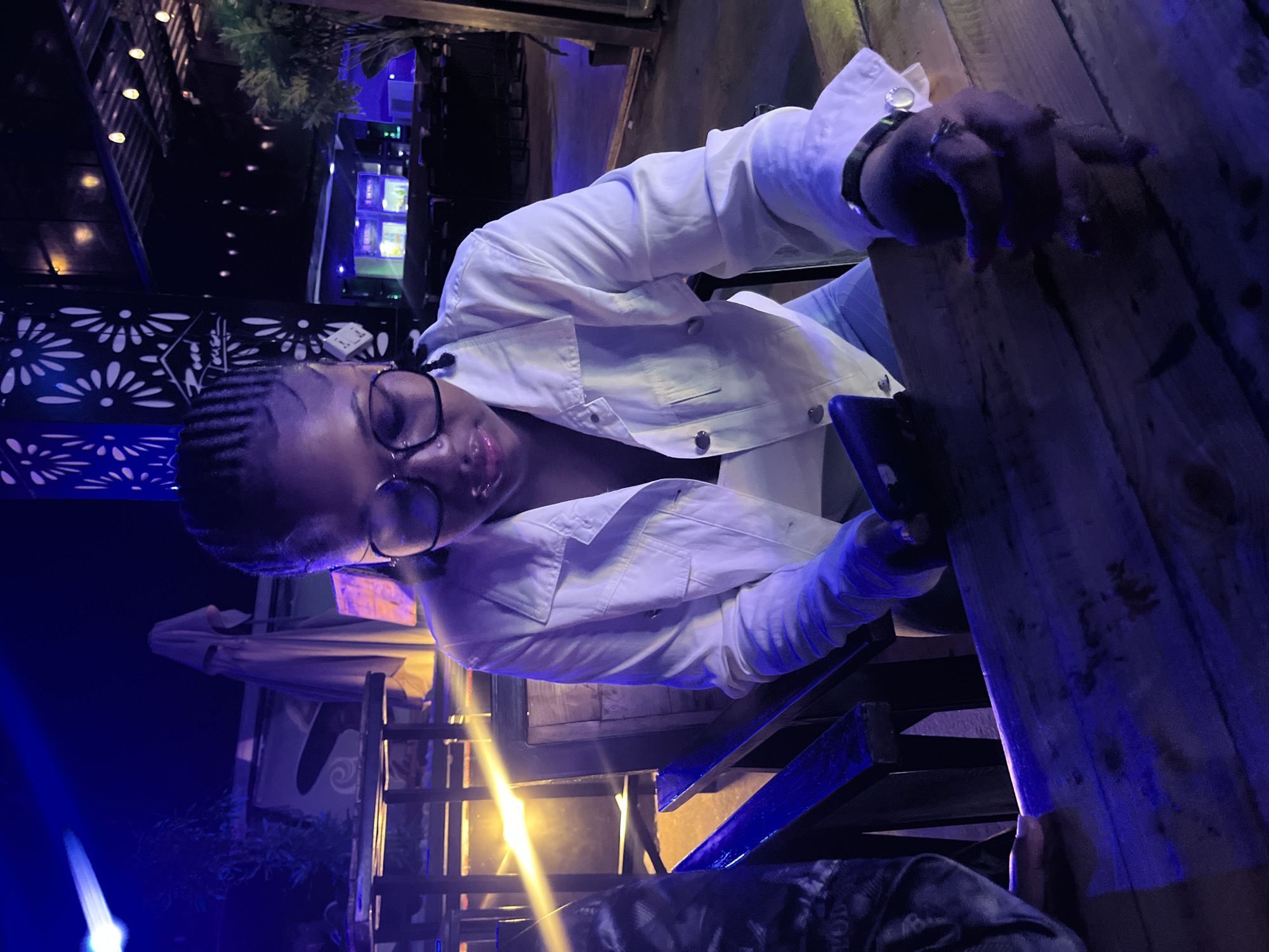 A young Black woman with braided hair and glasses is sitting at a wooden table, looking down at her phone. She is wearing a white jacket over a white top. The setting appears to be an outdoor or semi-outdoor restaurant or bar, with dim lighting and blue and yellow accents. In the background, there are tables, chairs, plants, and string lights. The overall atmosphere is casual and social. The location is a restaurant in the city of Ilorin, Nigeria.