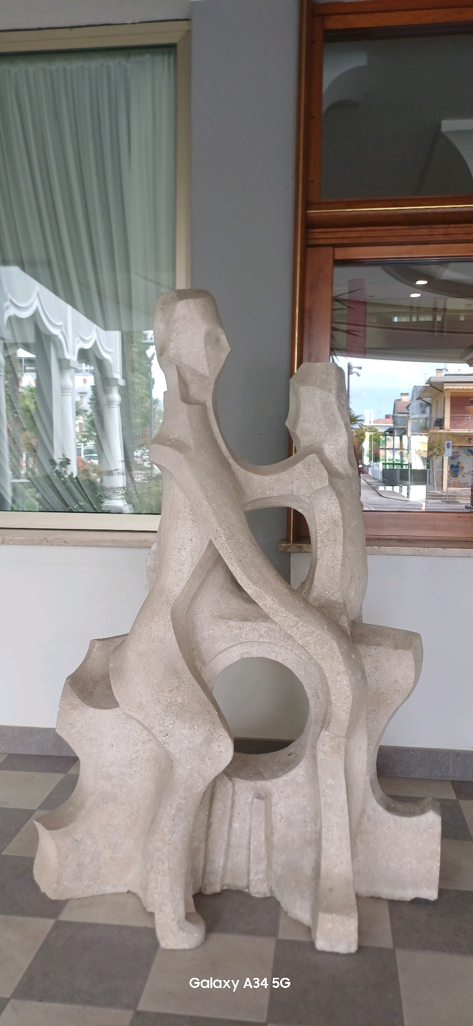 This image depicts a light-colored, abstract sculpture of two intertwined figures situated in the city of Abano Terme, Italy. The sculpture is made of a rough-textured material, possibly stone or concrete, and features rounded, flowing shapes with large openings. The figures appear to be seated or embracing.
The sculpture is placed on a checkered floor, with gray and beige squares. Behind the sculpture, there is a large window with pale green curtains visible through it, suggesting an interior space. To the right of the sculpture, a wooden-framed window looks out onto a street scene with buildings and trees. The architecture visible through the windows suggests a Mediterranean or European setting. The lighting is natural, with soft shadows indicating an overcast or diffused light source. The bottom right corner of the image displays the text "Galaxy A34 5G", indicating the device used for capture.
