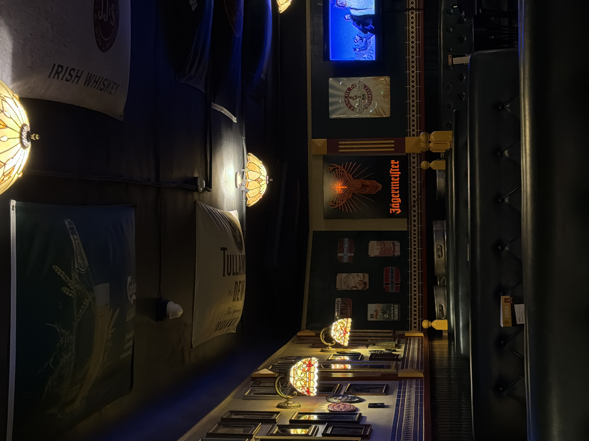 The image shows the interior of a pub or bar, potentially located in Rostov-on-Don, Russia, given the partially visible Russian text "ОБЕД." The interior features themed decorations including promotional banners for "Irish Whiskey" and "Tullamore Dew" along the walls. There are multiple lamps with stained-glass shades providing a warm, ambient light. The wall decor also includes framed pictures and advertising for "Jägermeister," contributing to a traditional pub atmosphere. A padded, upholstered seating area is visible along the right-hand side.