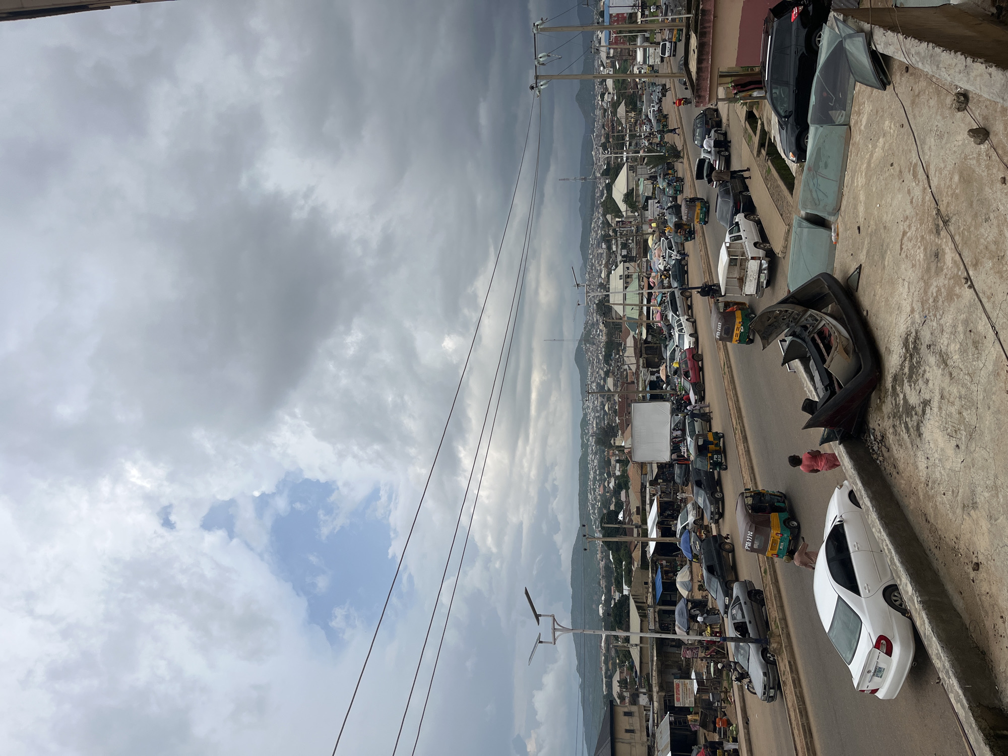 This is an eye-level, wide shot of a busy street in Rafin Par, Nigeria, under a cloudy sky. The scene is a mix of urban activity and a slight sense of disarray.
In the foreground, a concrete ledge is visible, with several car parts scattered on it, including what appears to be a bumper and some car doors. These parts are weathered and dusty, suggesting they are either discarded or awaiting repair.
The main focus of the image is the street itself, which is filled with traffic. Cars, taxis, and motorized rickshaws (known as Keke NAPEPs) are moving along the asphalt road. Some of the vehicles are white, silver, and black. There are also several people visible on the street and sidewalks, including pedestrians and individuals interacting with market stalls. The market stalls are set up along the side of the road, with various goods covered by umbrellas.
In the middle ground, buildings line the street, some appearing to be shops and residences. A white billboard is also visible. Further in the distance, the city stretches out with more buildings and a backdrop of rolling, green hills under a dramatic, overcast sky. Power lines crisscross the upper portion of the image, indicating the presence of infrastructure. The overall impression is one of a bustling urban environment, potentially facing a period of overcast weather.