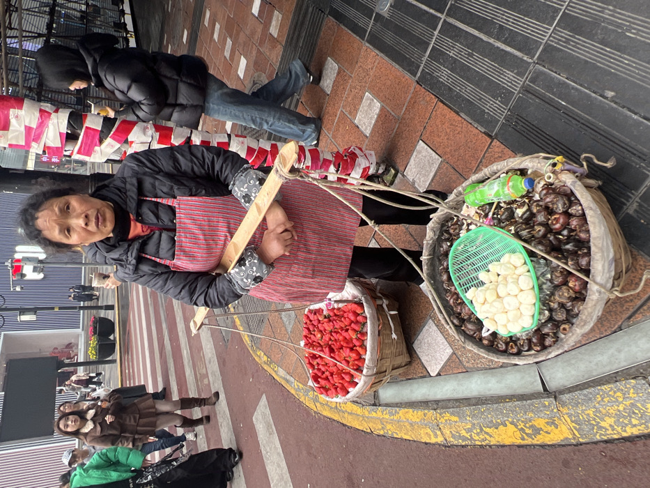 An Asian woman wears an apron and carries two baskets suspended from a wooden yoke. One basket holds bright red strawberries, while the larger basket contains brown bulbs and a green basket filled with peeled white bulbs. The vendor stands on a street corner near a crosswalk where people walk by. A person in a puffy jacket and jeans is partially visible in the background. The ground is paved with red and gray tiles and has a yellow curb.