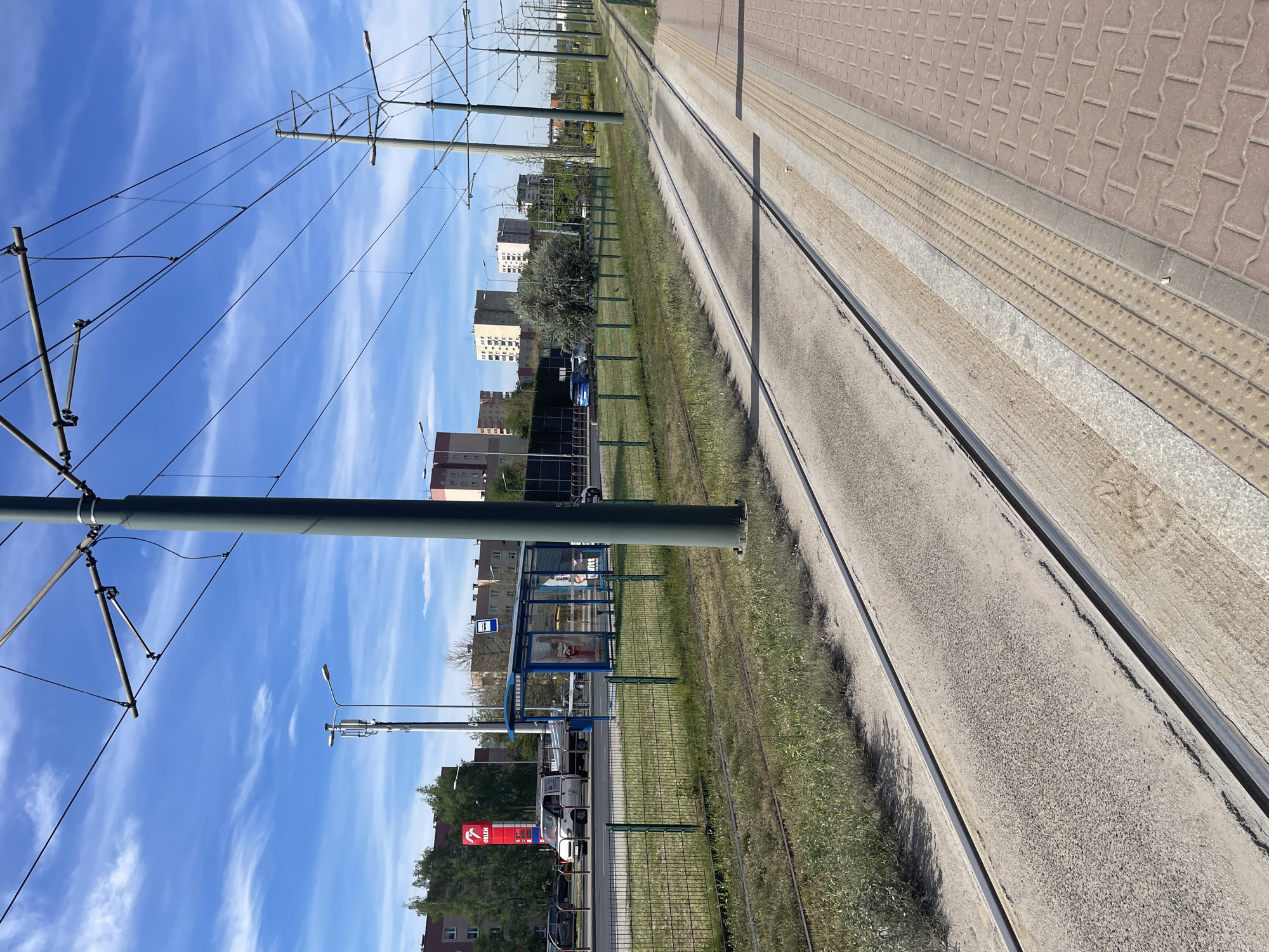 Here is a description of the image:
The image shows a street-level view of a tram line with a tram stop in an urban setting. The tram tracks run parallel to a pedestrian walkway, which is paved with a combination of brick-like tiles and tactile paving. A green fence separates the tram tracks from the adjacent road. Overhead tram wires are suspended from metal poles. In the background, there are several buildings, including apartment blocks. A blue tram stop shelter is visible, and a gas station with a red logo can be seen across the street. The sky is blue with scattered clouds.
