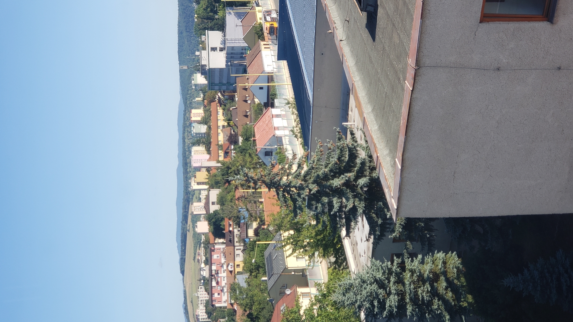 The image is an elevated, clear daytime, outdoor shot looking out over a cityscape. The sky is a bright, cloudless blue. In the foreground, a grey, textured building with a flat, dark roof occupies the lower right and bottom of the frame. A thin copper trim runs along the edge of this roof. Two dark green coniferous trees are visible on the left side of the foreground, with one partially obscuring a portion of the grey building.
In the middle ground, several residential houses with tiled roofs are nestled amongst mature green trees. Some of these houses have light-colored walls, while others are a pale yellow. A few of these houses are bordered by a light-colored fence. Further back, a street is visible with a yellow truck parked near a building.
The background features a line of multi-story apartment buildings, predominantly in light colors, stretching across the horizon. Some of these buildings are tall and slender, while others are shorter and wider. To the far right of the background, more distant trees and rolling hills create a natural backdrop for the urban environment. The overall scene depicts a residential area within a city.
This image is a view of the Šarišská vrchovina region, specifically from the Sídlisko Sekčov area in Prešov, Slovakia, looking towards the older part of the city and the distant hills.