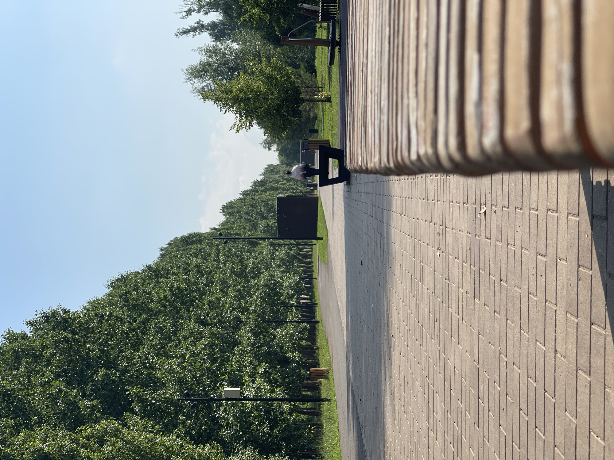 The image depicts a paved pathway in a park-like setting. On the left, a dense row of tall, leafy green trees lines the path, with streetlights visible between them. The pathway itself is paved with rectangular bricks, laid in a pattern. In the middle ground, a dark-colored bench is occupied by a person facing away from the camera, wearing a light-colored top. Beyond the bench, the park continues with more trees and green grass, and some outdoor exercise equipment can be seen to the right. The sky above is clear and blue with a few faint clouds. The foreground is dominated by the textured surface of the paved area and a close-up view of a wooden bench. Based on the surrounding context and typical park infrastructure, this scene is likely located in a public park in the city of Orenburg, Russia.