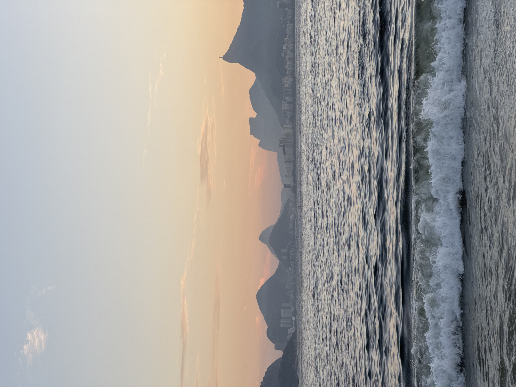 The image captures a panoramic view of Rio de Janeiro's coastline from Niterói, Brazil, at dusk. The sky is a soft gradient of pale blue and warm peach, with wisps of clouds. In the distance, silhouetted against the sky, are several iconic mountains, including Corcovado with the Christ the Redeemer statue visible atop its peak, and the Dois Irmãos mountains. A city skyline of buildings is nestled at the base of these mountains. The foreground is dominated by the gentle waves of the Guanabara Bay, with a foamy wave cresting closer to the viewer. The water is dark and reflective, mirroring the muted colors of the sky and the distant landscape. No people or animals are visible in the frame.